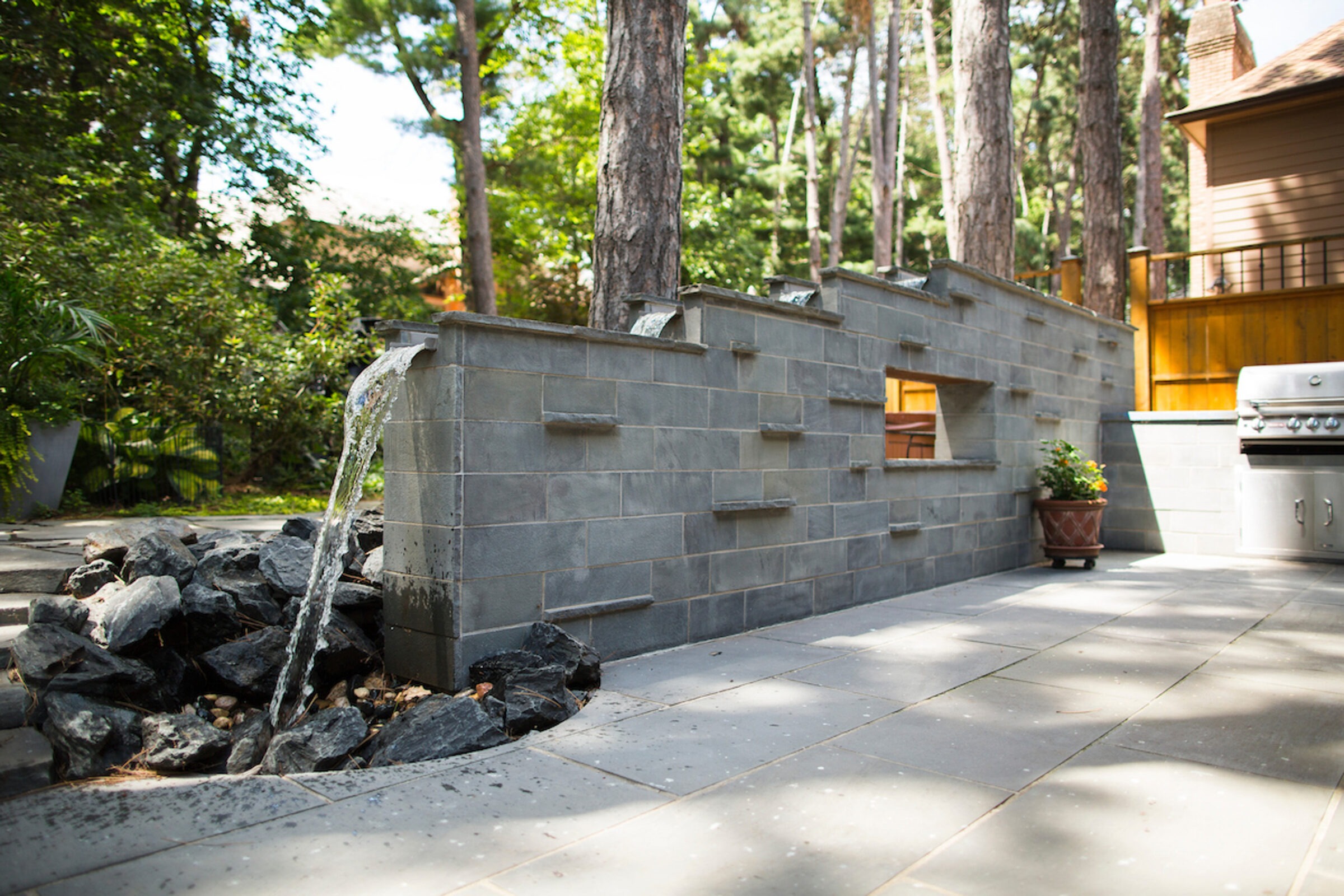 Outdoor patio with a stone water feature, barbecue grill, and potted plants surrounded by trees, under soft sunlight.