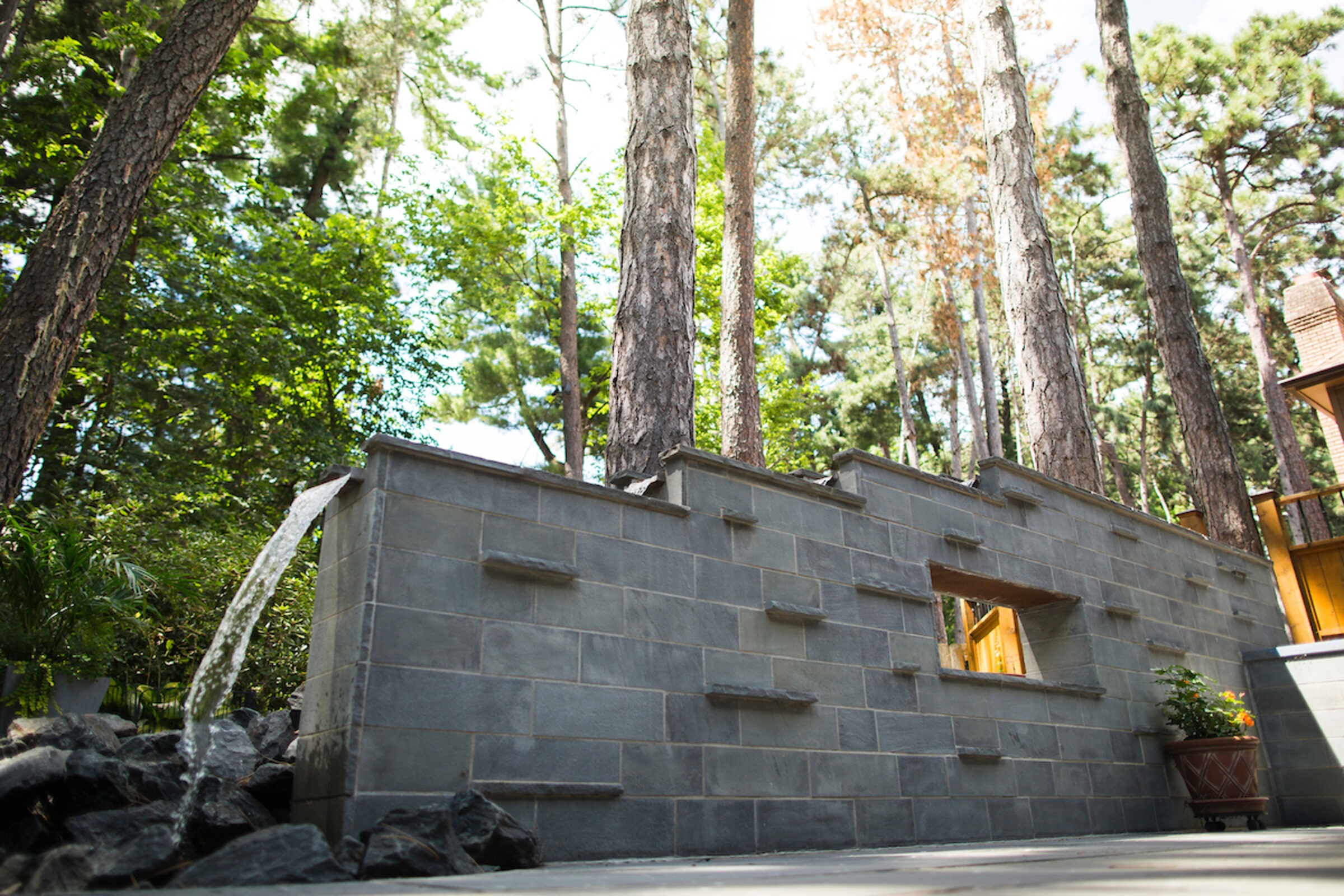 A modern outdoor water feature flows from a brick wall surrounded by trees and plants, creating a serene natural setting.