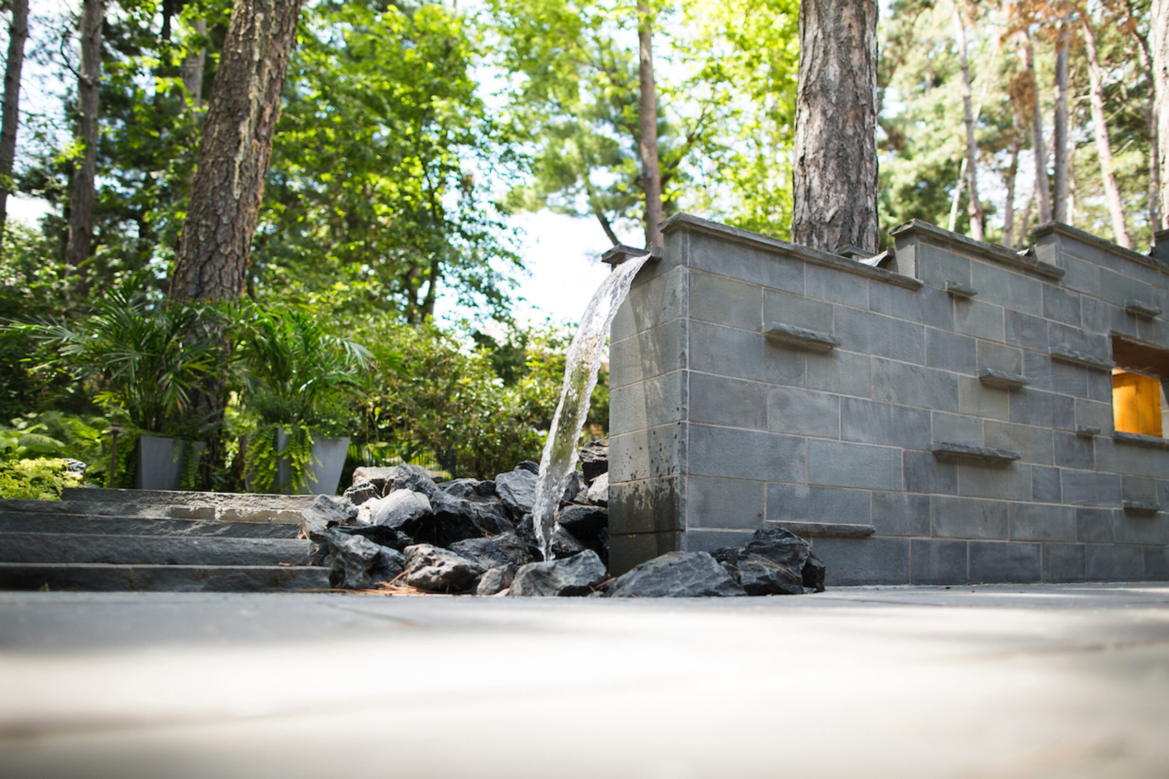 A serene outdoor scene featuring a small stone fountain with flowing water, surrounded by rocks, trees, and lush greenery.