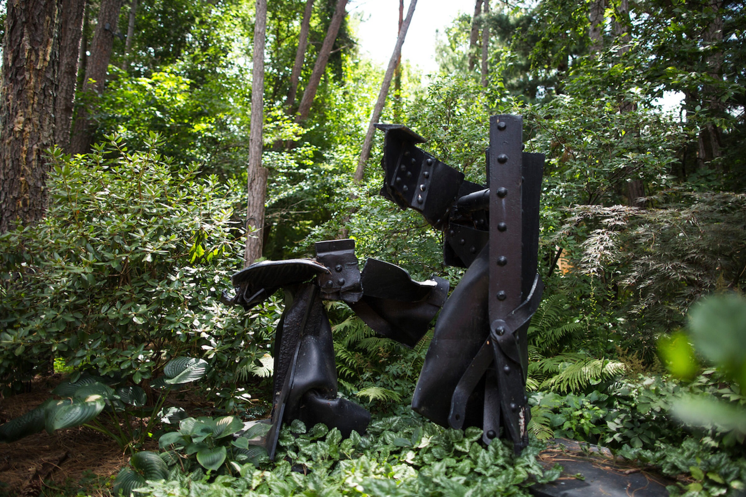 Abstract black metal sculpture set amidst lush green forest, surrounded by trees and dense foliage, creating a serene natural backdrop.