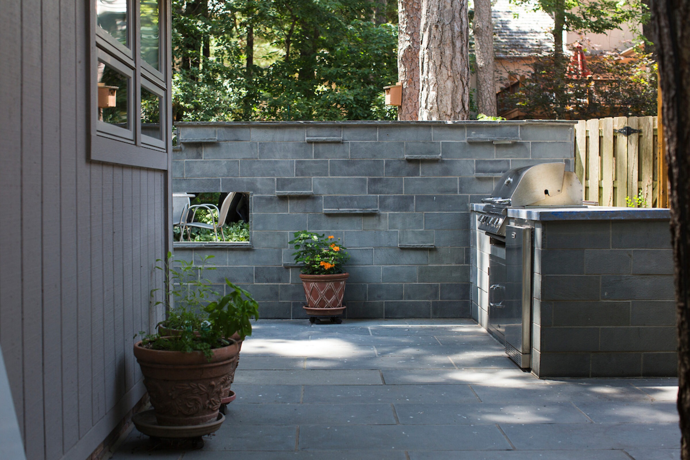 A backyard patio with potted plants, a stone wall, and a built-in grill. Surrounding greenery suggests a tranquil outdoor setting.