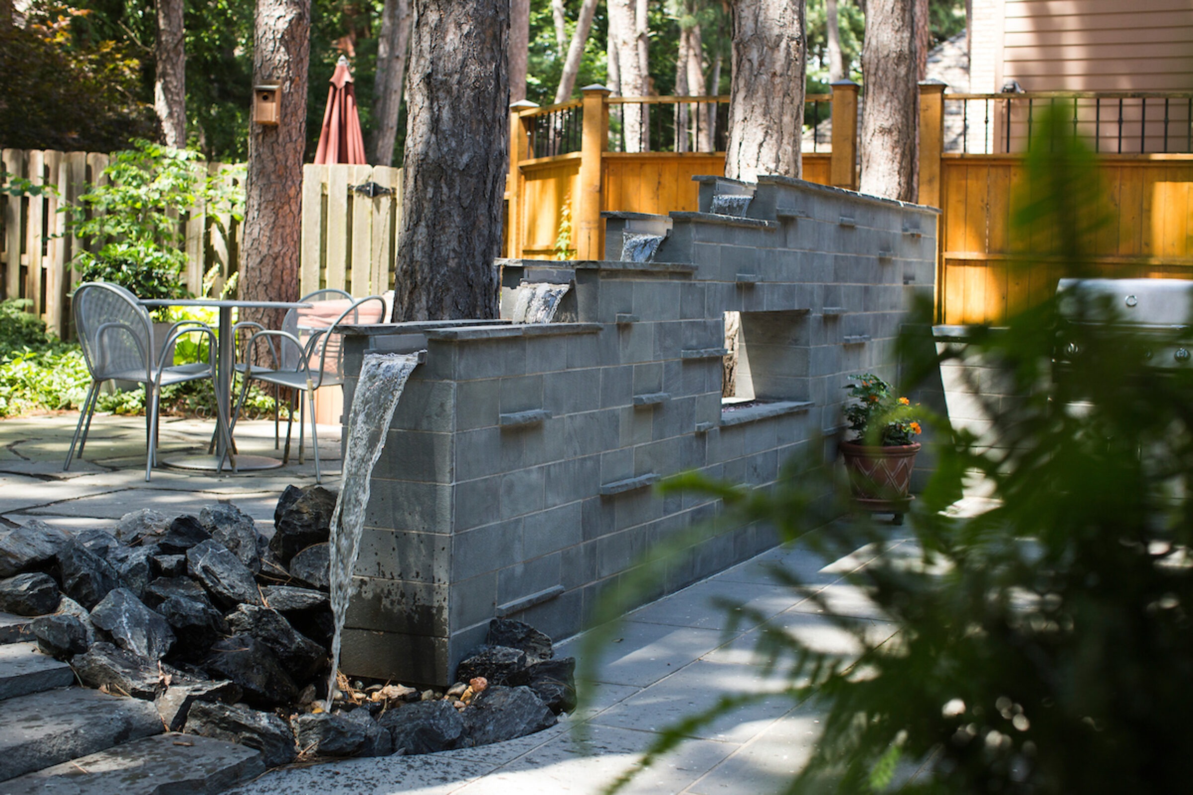 A serene backyard with a tiered stone water feature, patio furniture, and lush greenery, creating a peaceful outdoor retreat.