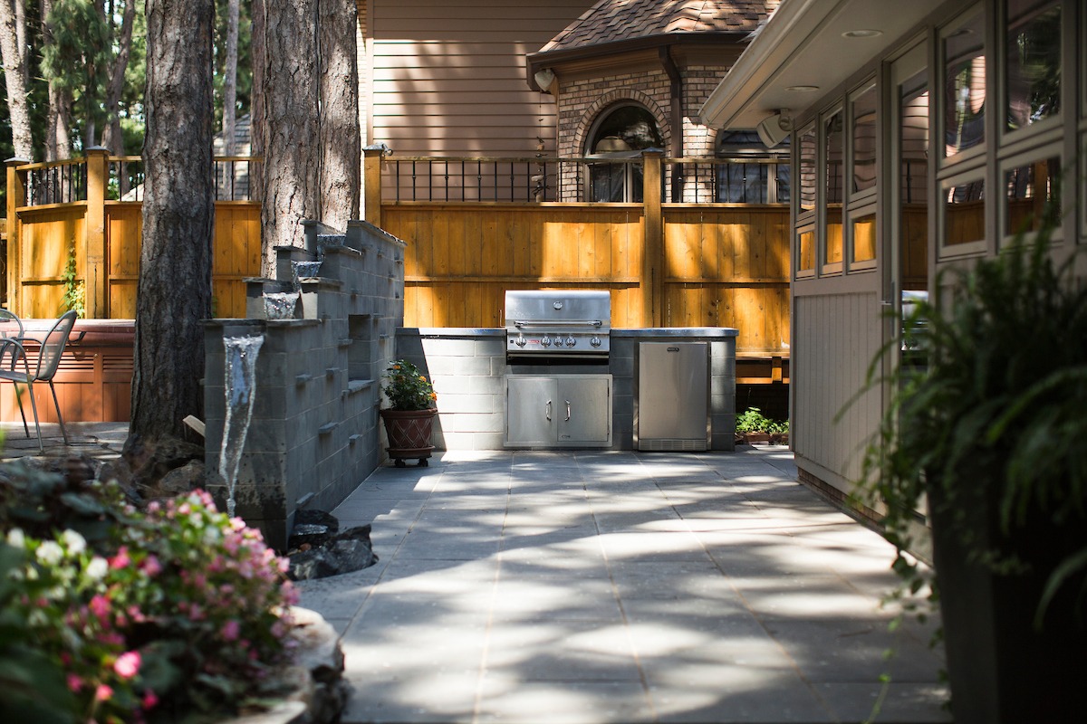 Outdoor patio with stainless steel grill, stone floor, and flowers beside wooden fence. Trees provide shade; house visible in background.