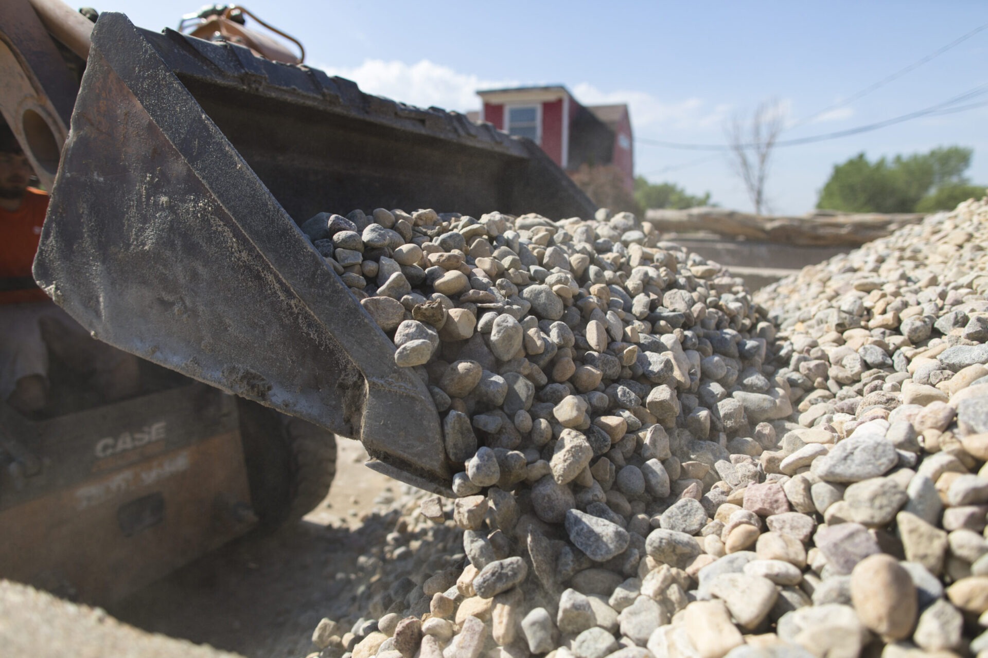 A construction vehicle's bucket unloads gravel. A person operates the vehicle in an outdoor setting with a building visible in the background.