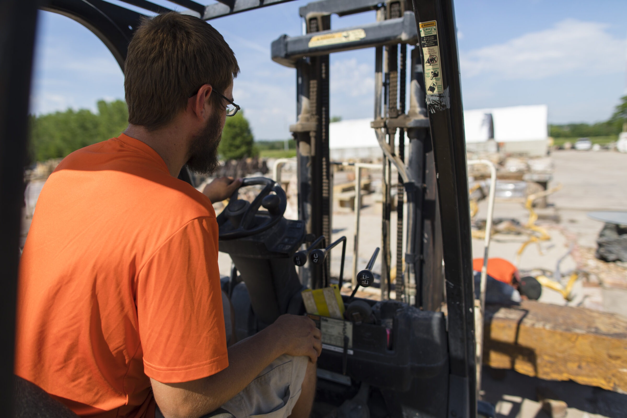A person operates a forklift outdoors, wearing an orange shirt. Another person is seen in the background working near wooden pallets.