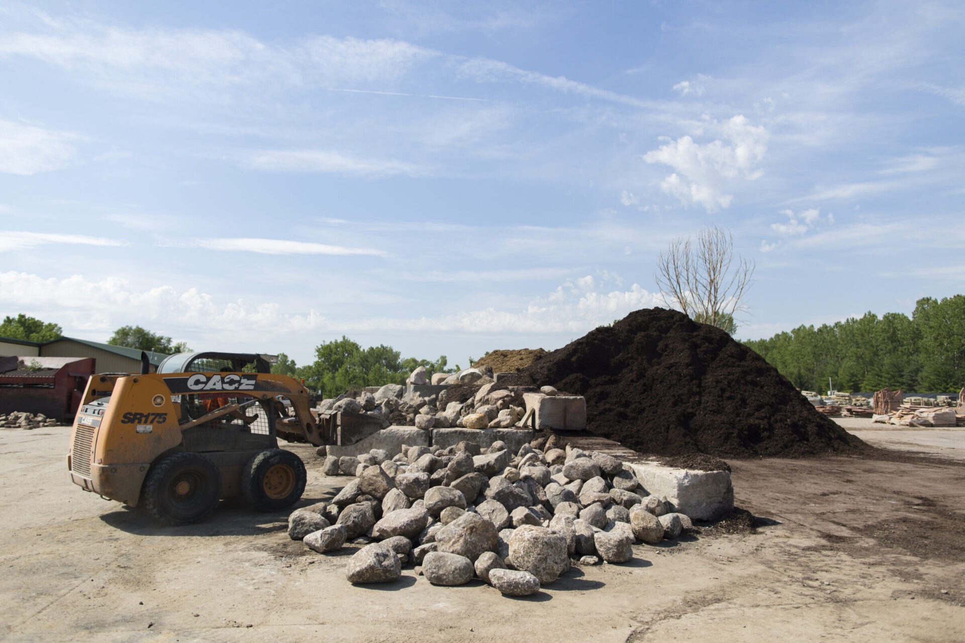 A construction site features a skid steer loader, rocks, and a soil pile under a clear blue sky, surrounded by green trees.