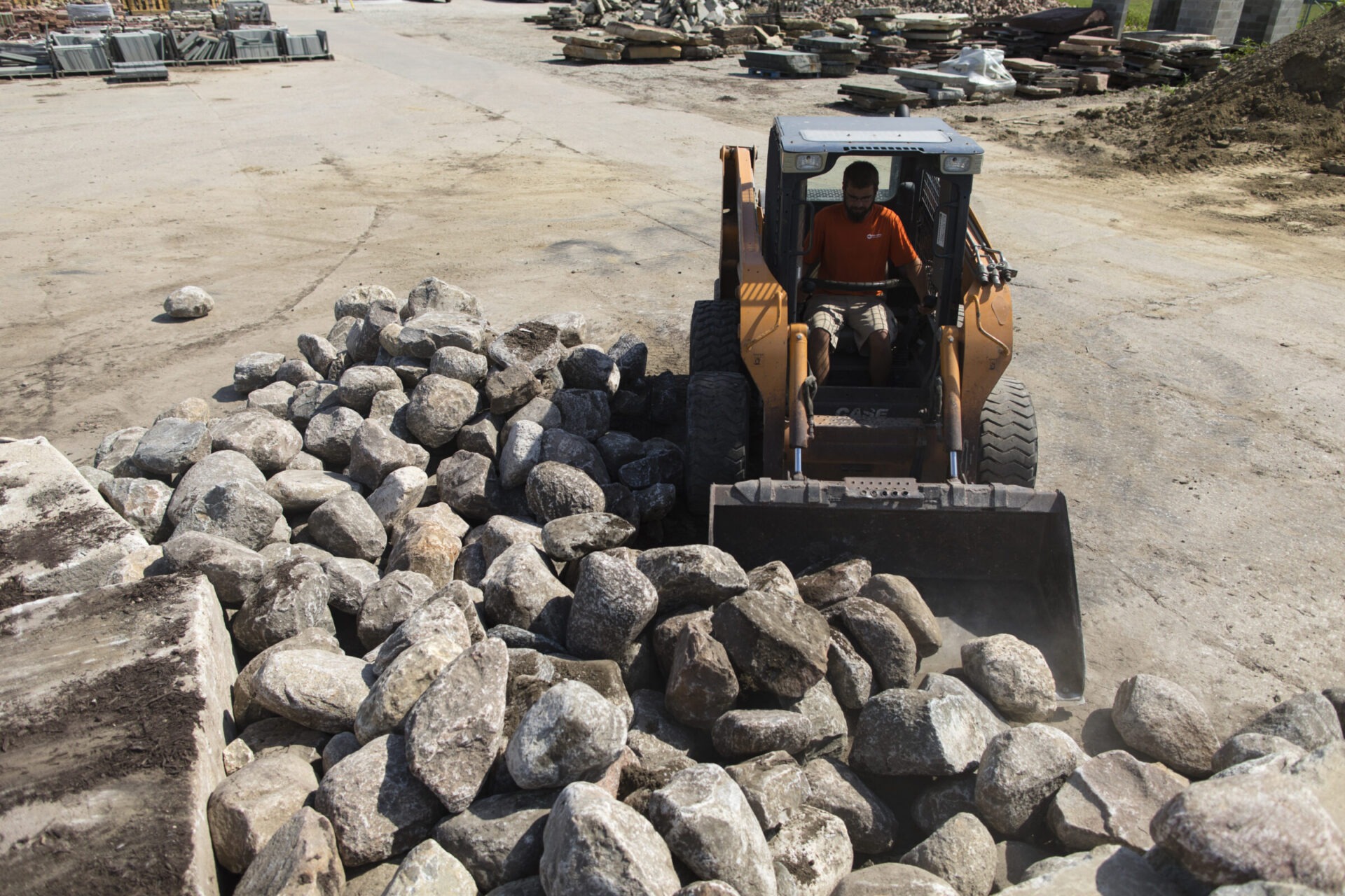 A person operates a skid steer loader moving large rocks in an outdoor construction site, surrounded by piles of materials and dirt.