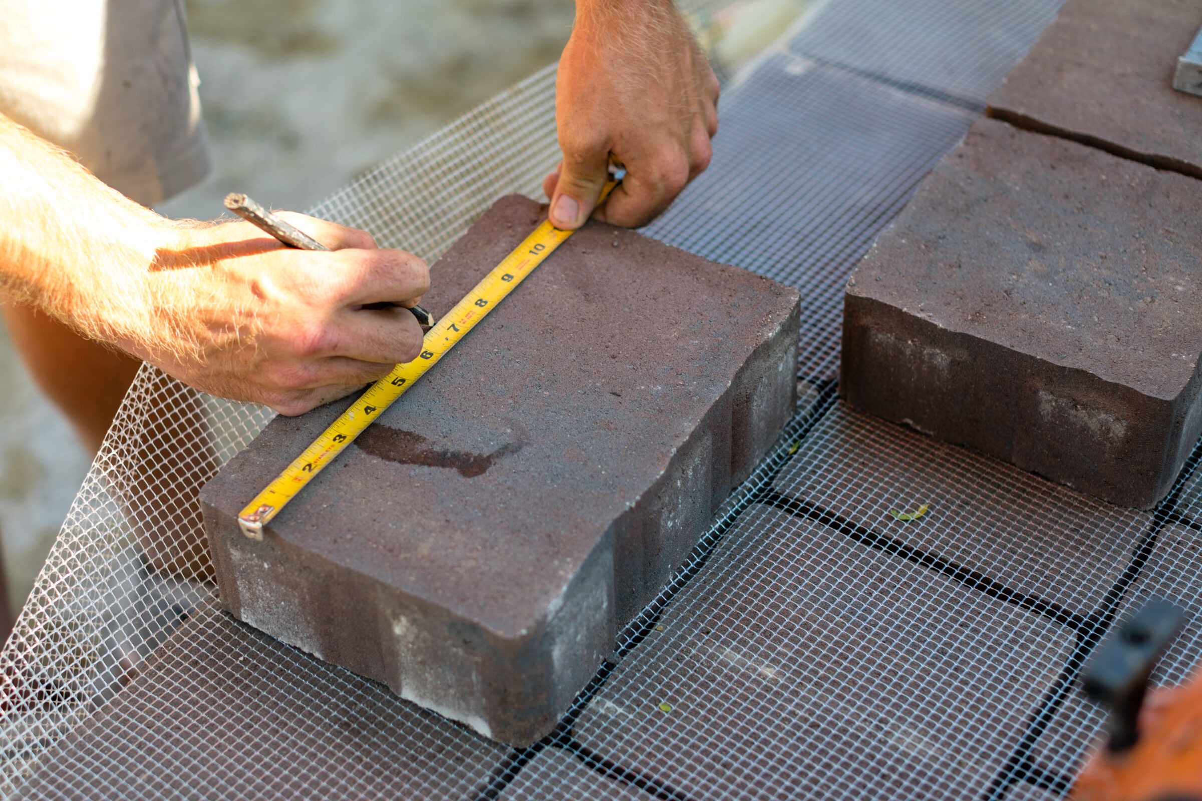 A person uses a measuring tape and pencil to mark dimensions on a brick placed on a wire mesh surface.