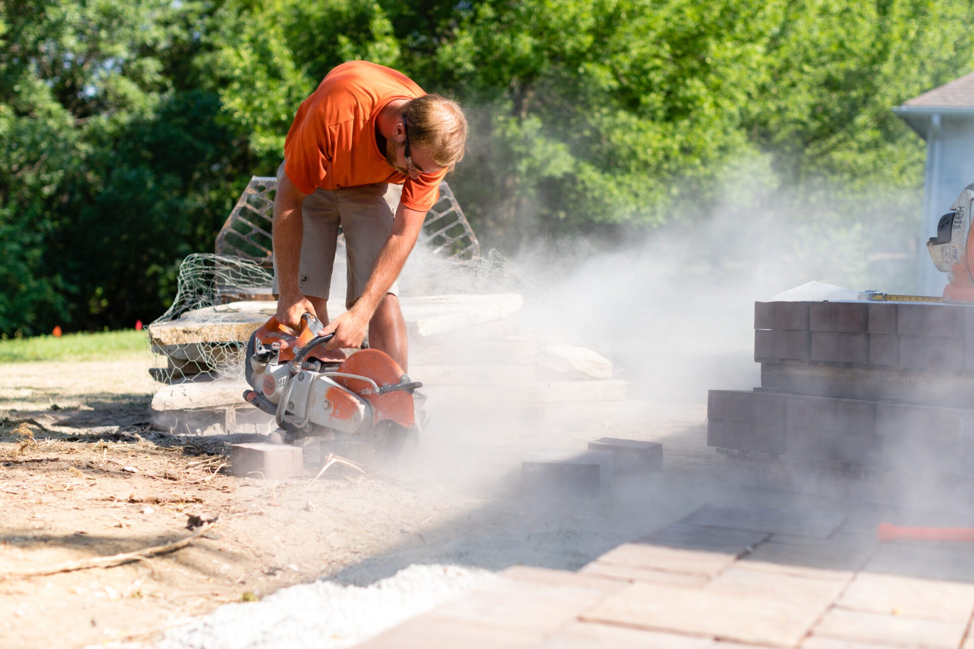 A person uses a power saw to cut bricks, generating dust. They're outdoors, surrounded by trees and construction materials.