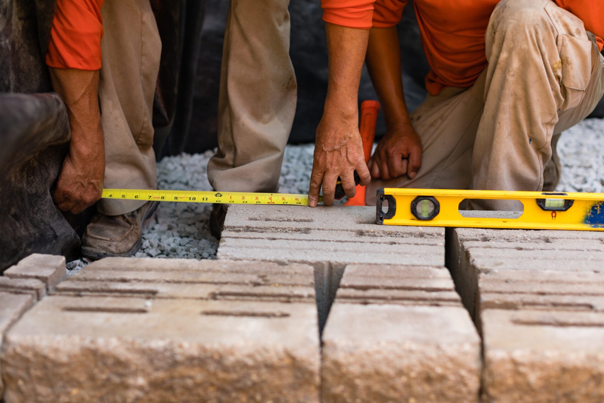 Two people measuring and leveling interlocking concrete blocks on gravel, using a tape measure and spirit level, wearing orange shirts and khaki pants.