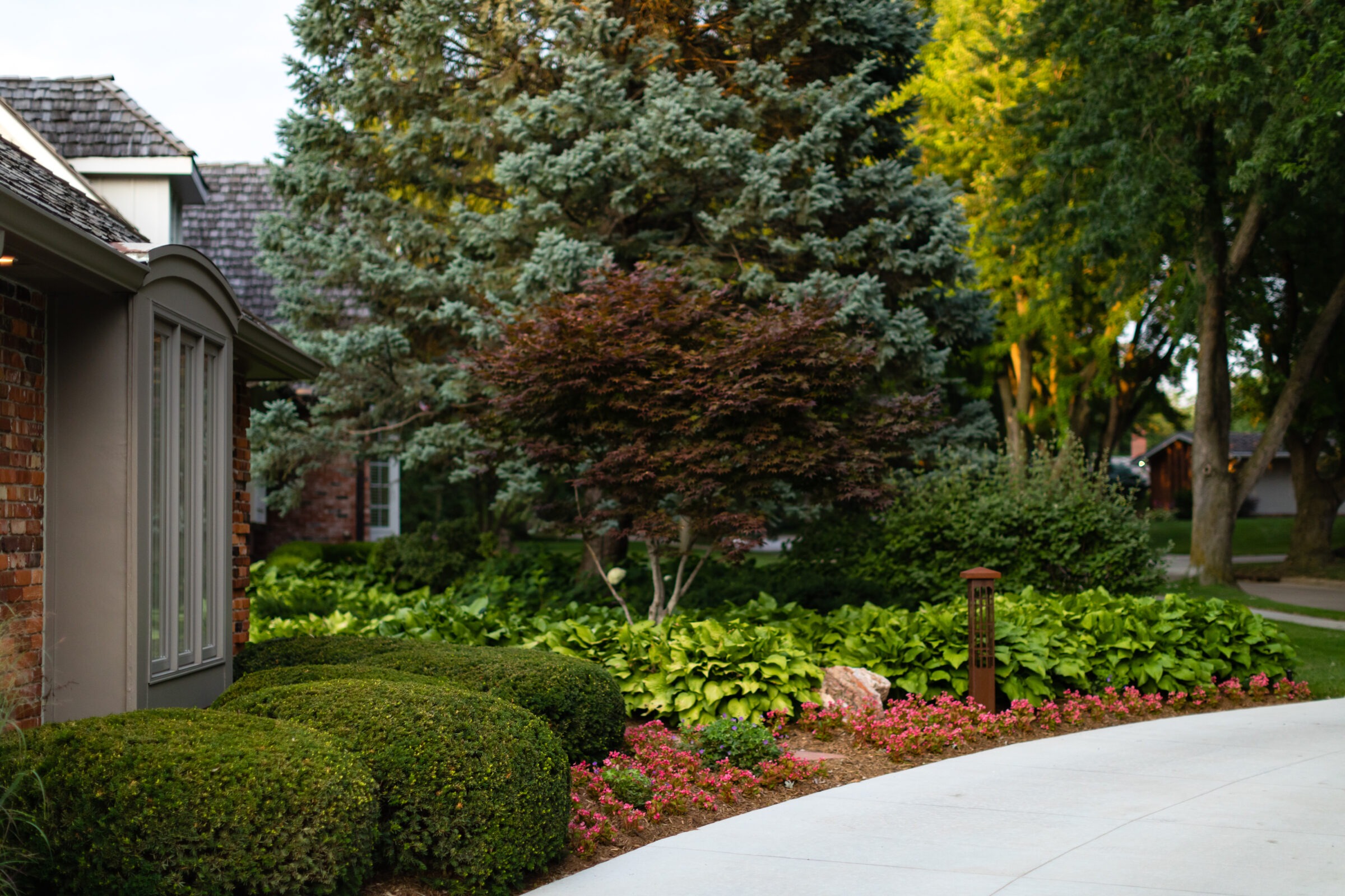 A beautifully landscaped garden with lush greenery and well-maintained shrubs surrounds a brick house with a shingled roof, at sunset.