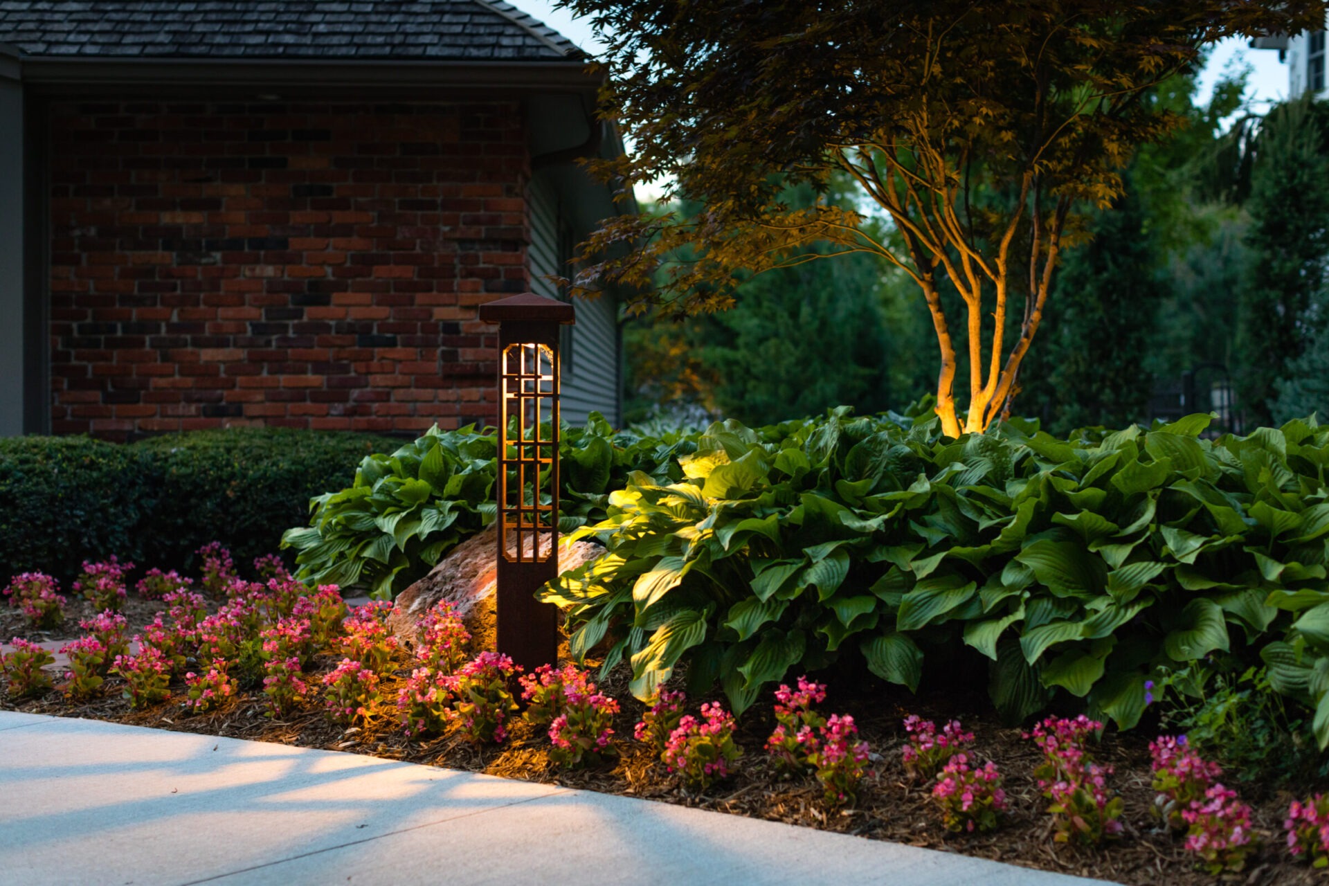 A garden scene with a lit lantern, vibrant pink flowers, lush green foliage, and a brick building backdrop at dusk.