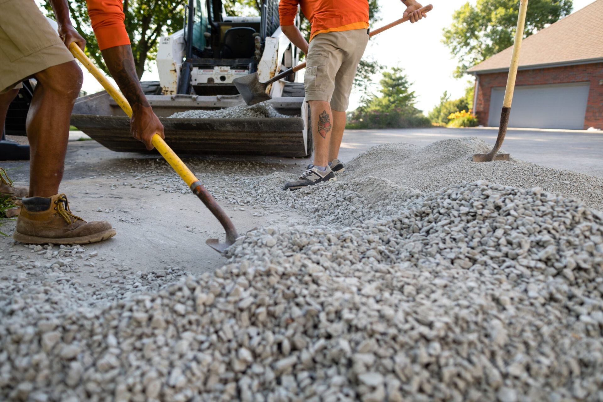 Two persons in casual attire using shovels to spread gravel from a pile near an excavator. There's a brick building and trees nearby.