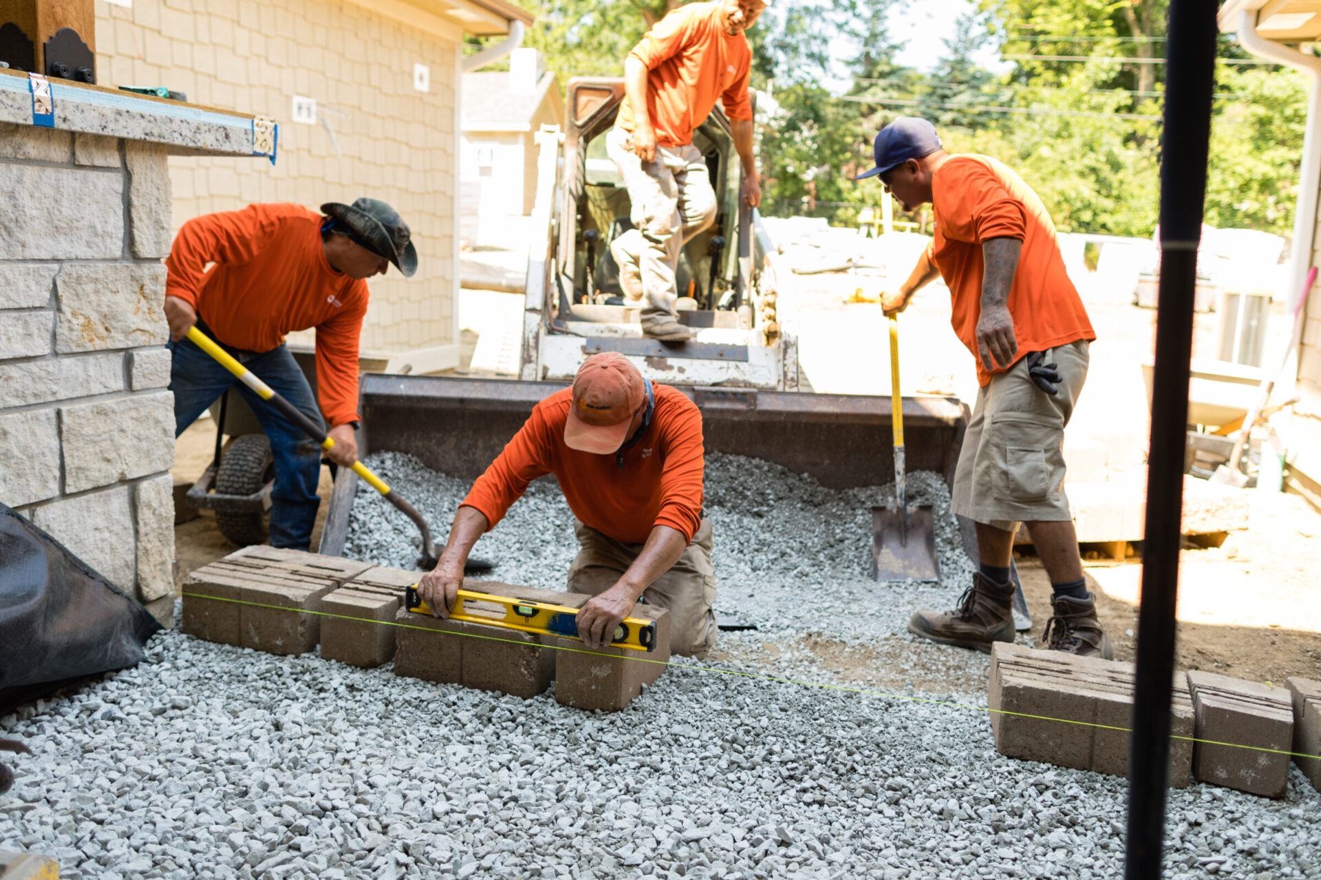 Four people in orange shirts work on a construction project, positioning bricks and shoveling gravel near a house.