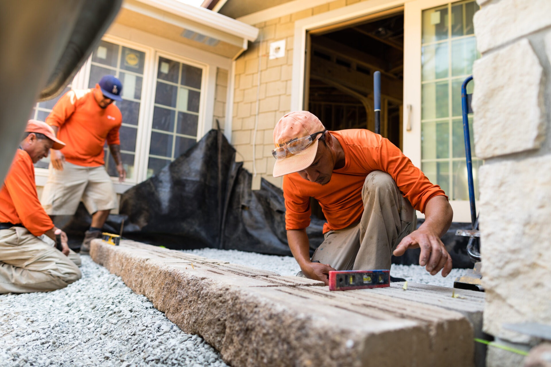 Three people in orange shirts work on a construction project, measuring cement blocks in front of a building with tan siding and windows.