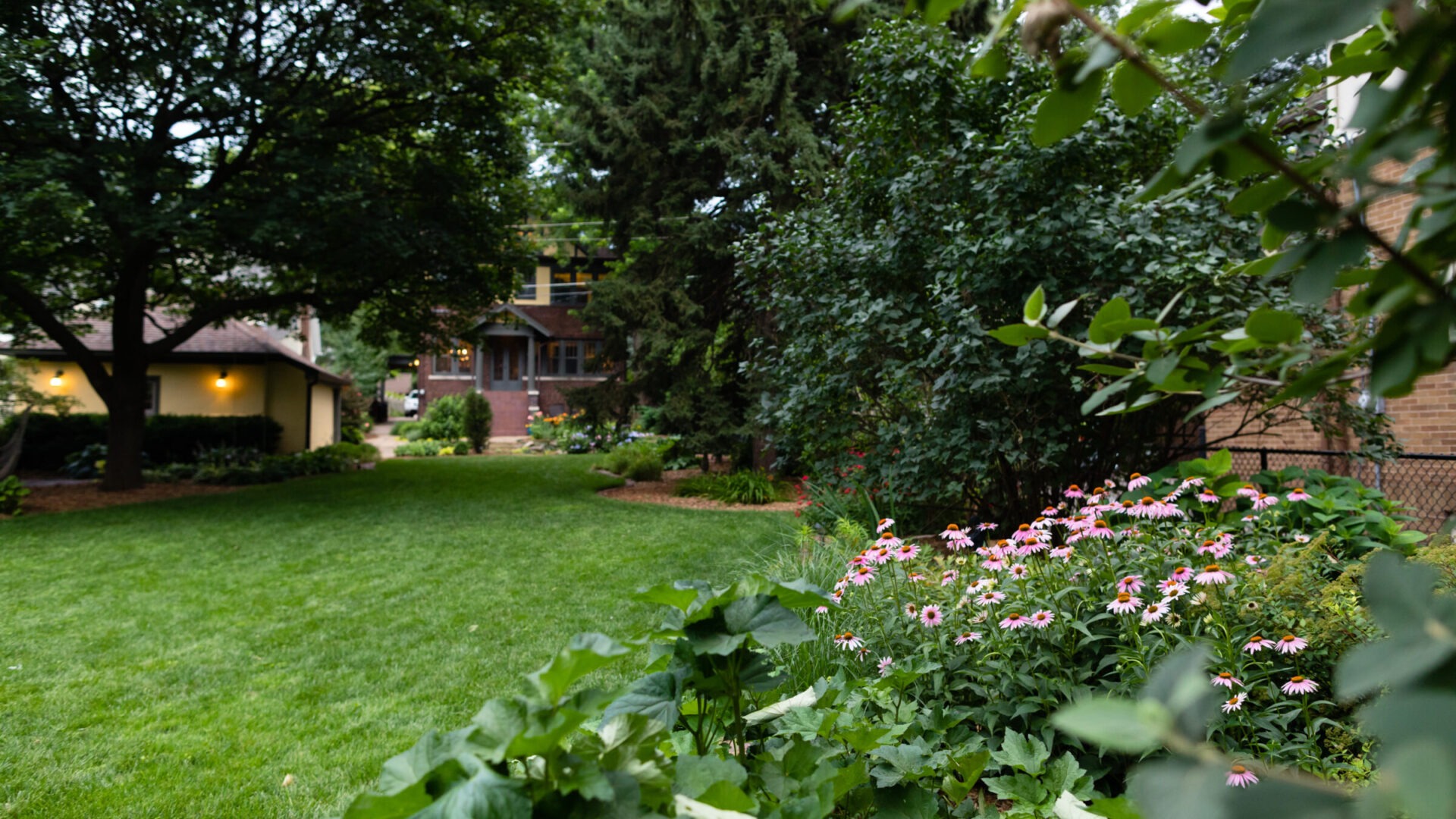 A lush garden landscape with blooming flowers, large trees, and a pathway leading to a cozy brick house surrounded by greenery.