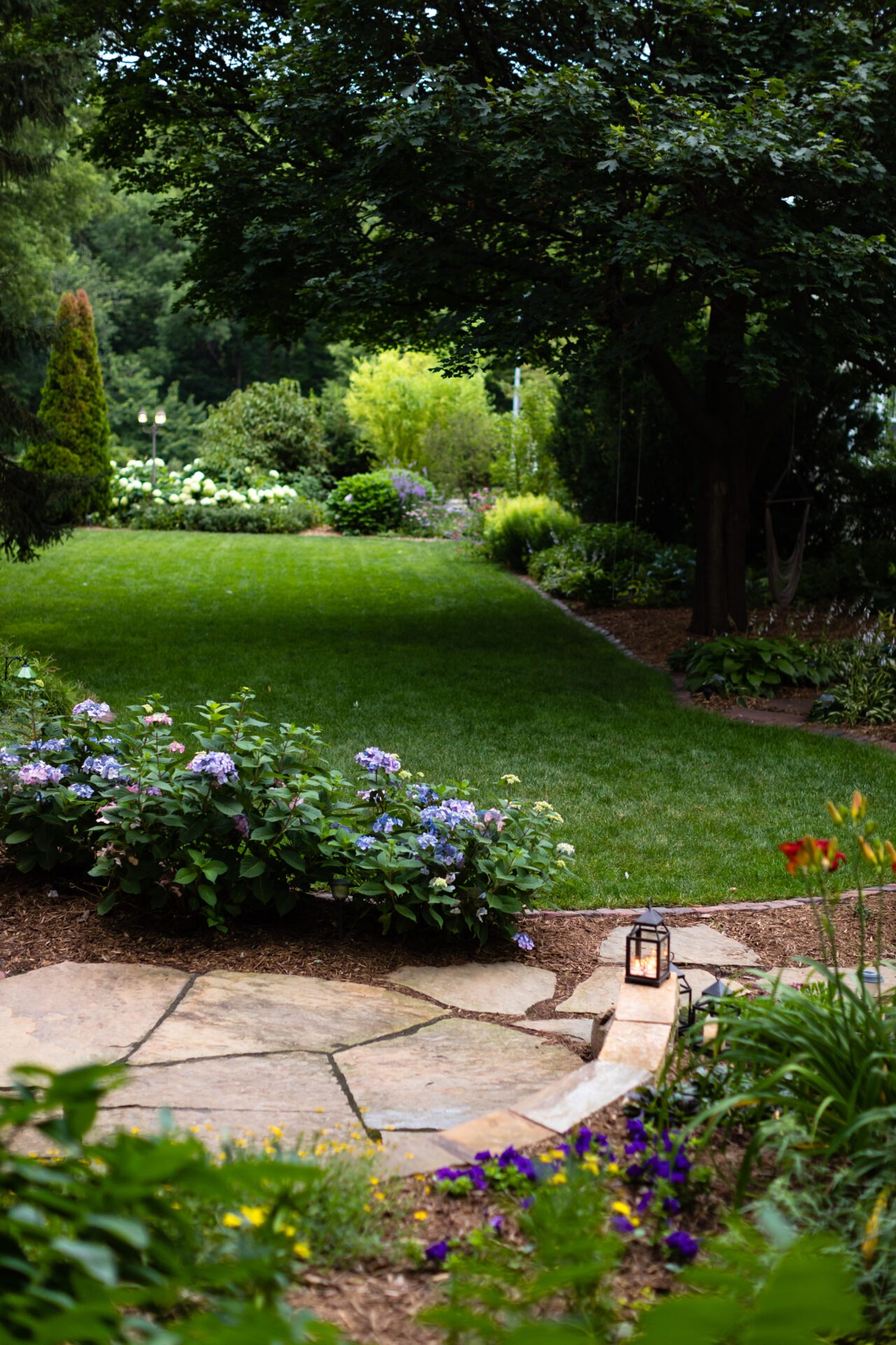 A lush garden scene features vibrant flowers, a well-manicured lawn, and a stone pathway illuminated by a small lantern.