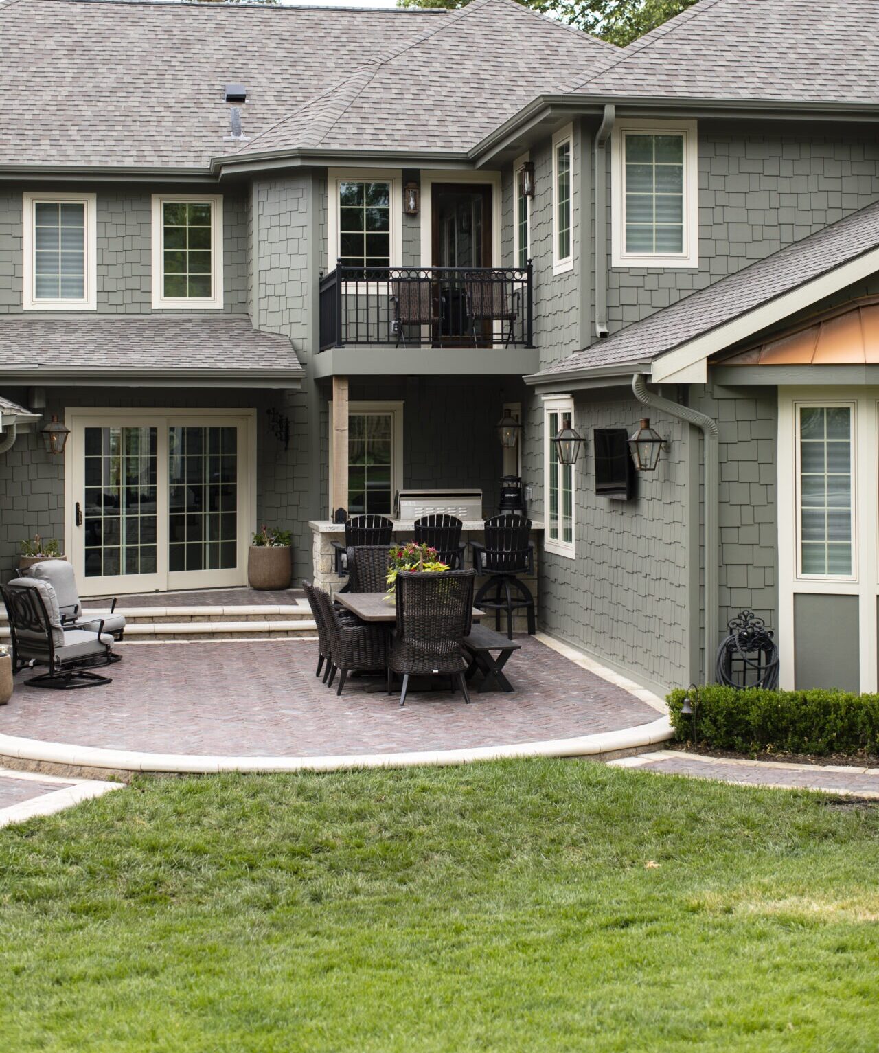 A gray house showing off one of our clients' outdoor kitchens by a house with shingle siding, featuring a patio with outdoor furniture and potted plants. Trees surround the property in the background.