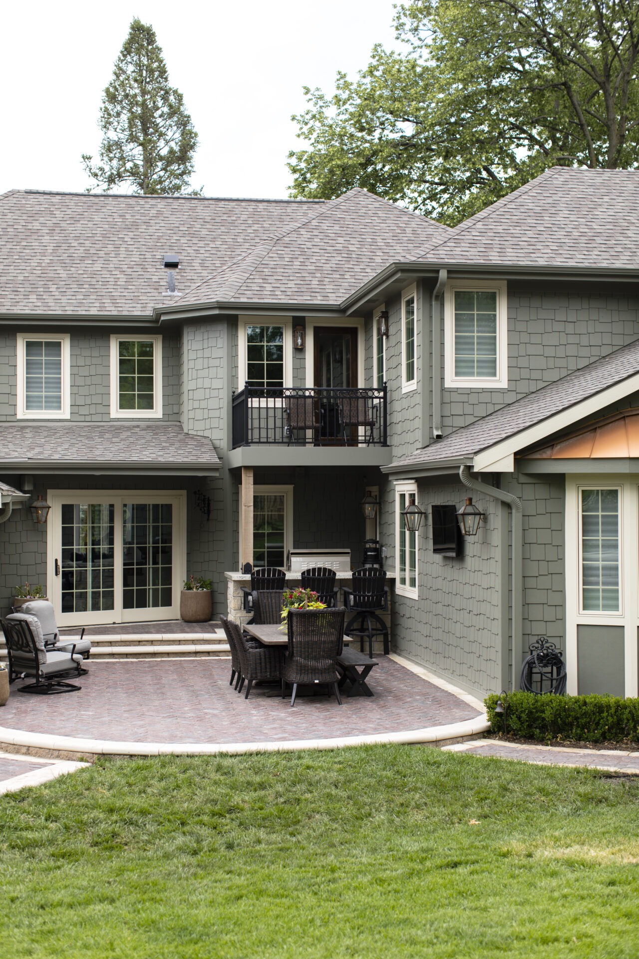 A gray house with shingle siding, featuring a patio with outdoor furniture and potted plants. Trees surround the property in the background.