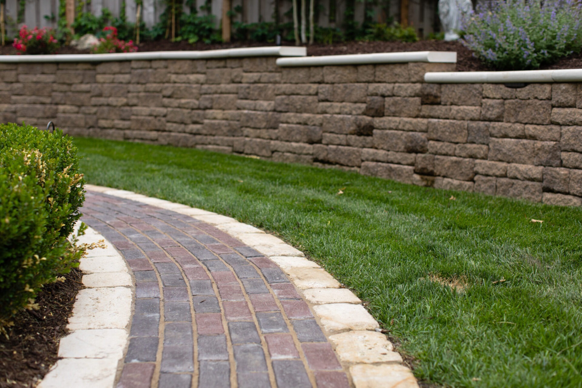 Curved brick path bordered by greenery leads along a manicured lawn, with a stone retaining wall in the background.