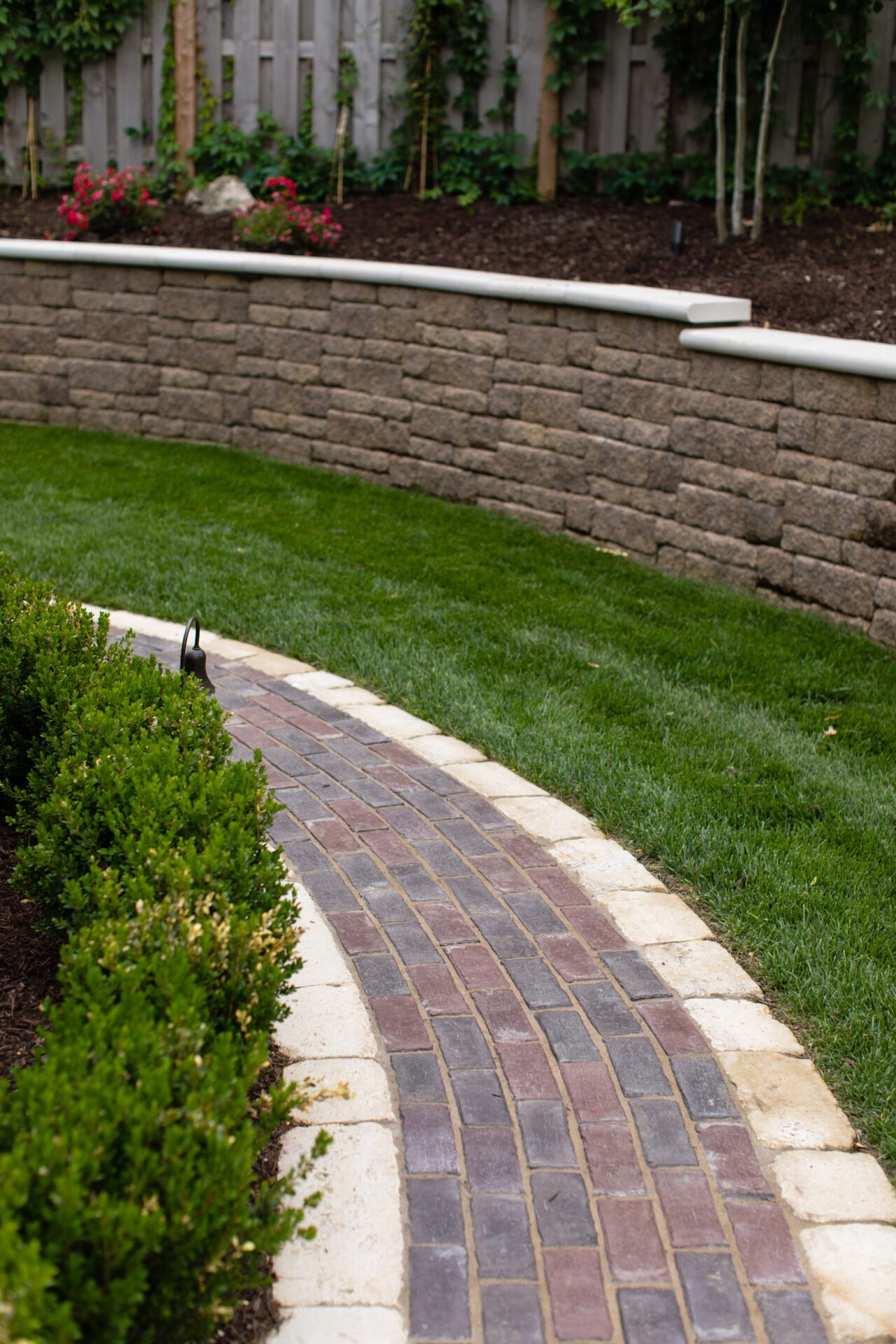 Brick pathway curves between neatly trimmed bushes and grass, leading to a stone retaining wall and wooden fence, with small plants visible.