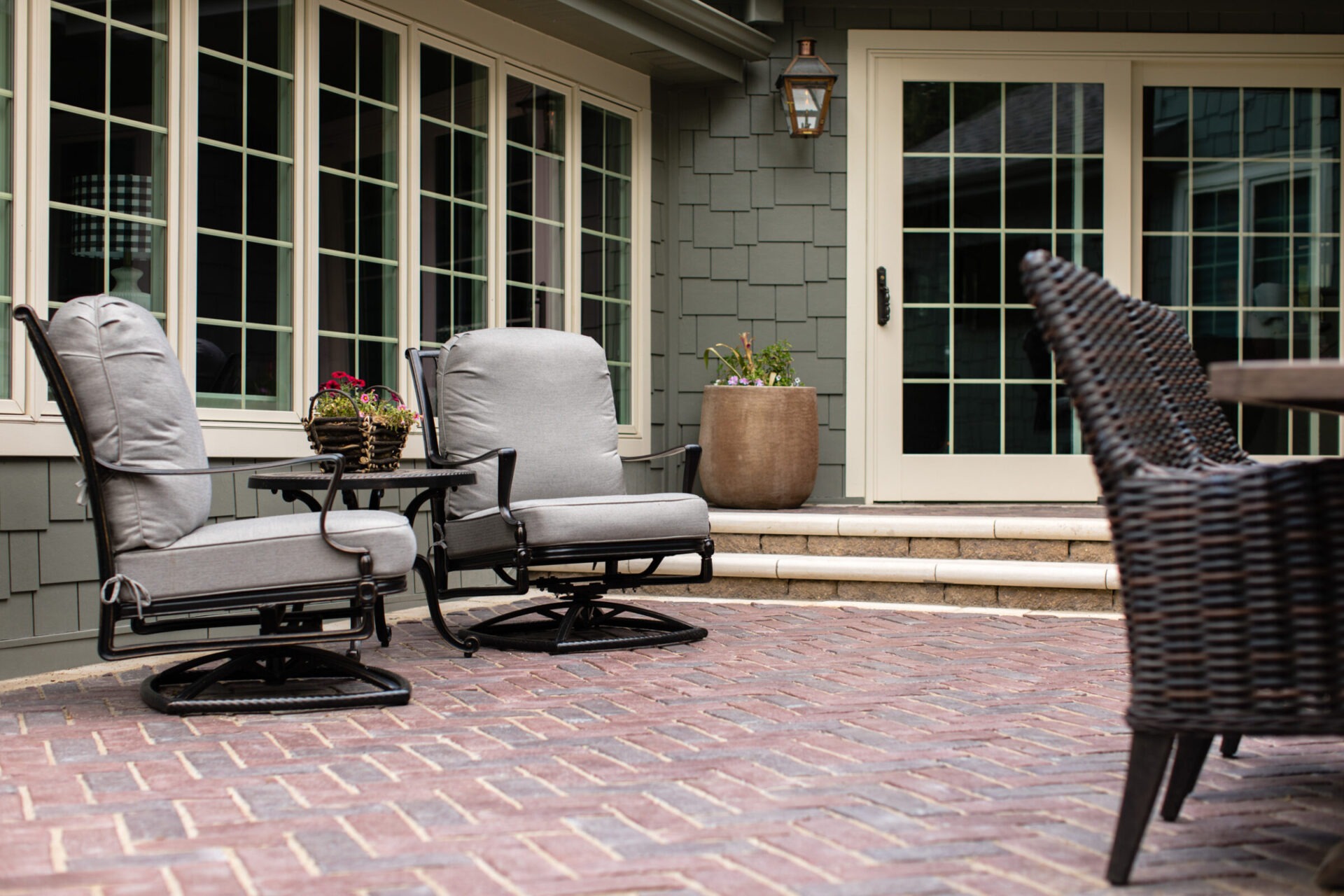 A cozy patio with cushioned chairs, wicker furniture, a brick floor, and potted flowers, next to a house with a large windowed door.