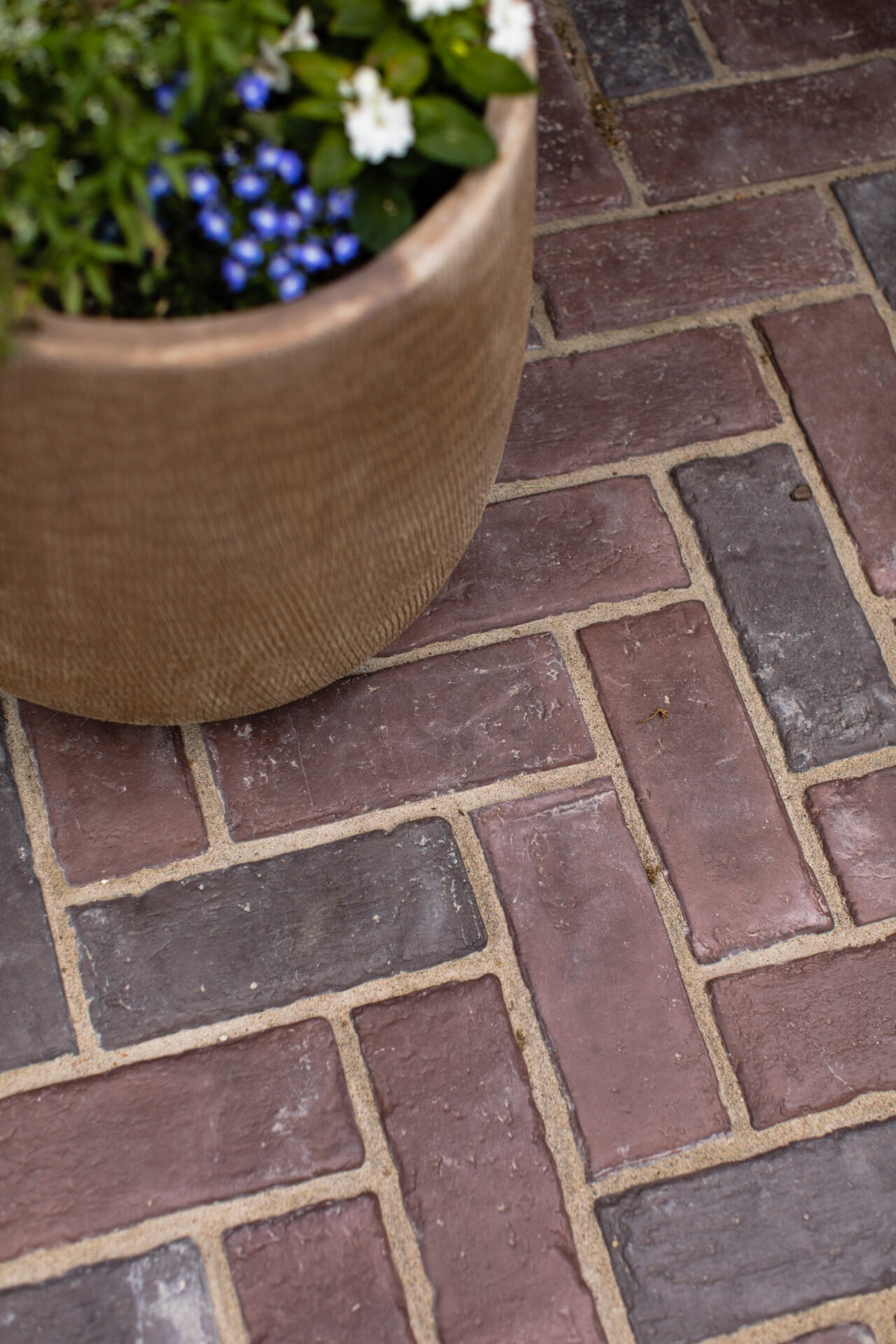 A large planter with blue and white flowers sits on a herringbone-patterned brick pavement, surrounded by green foliage.