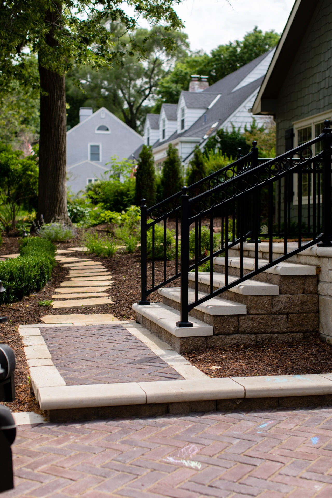 Stepped walkway with black railing leads through a landscaped garden, lined with hedges and trees, towards houses in a verdant neighborhood.