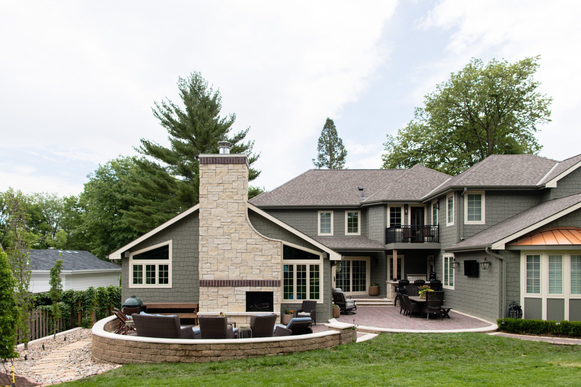 Spacious modern house with large patio, stone fireplace, outdoor seating, and surrounding trees, creating a serene and inviting atmosphere.