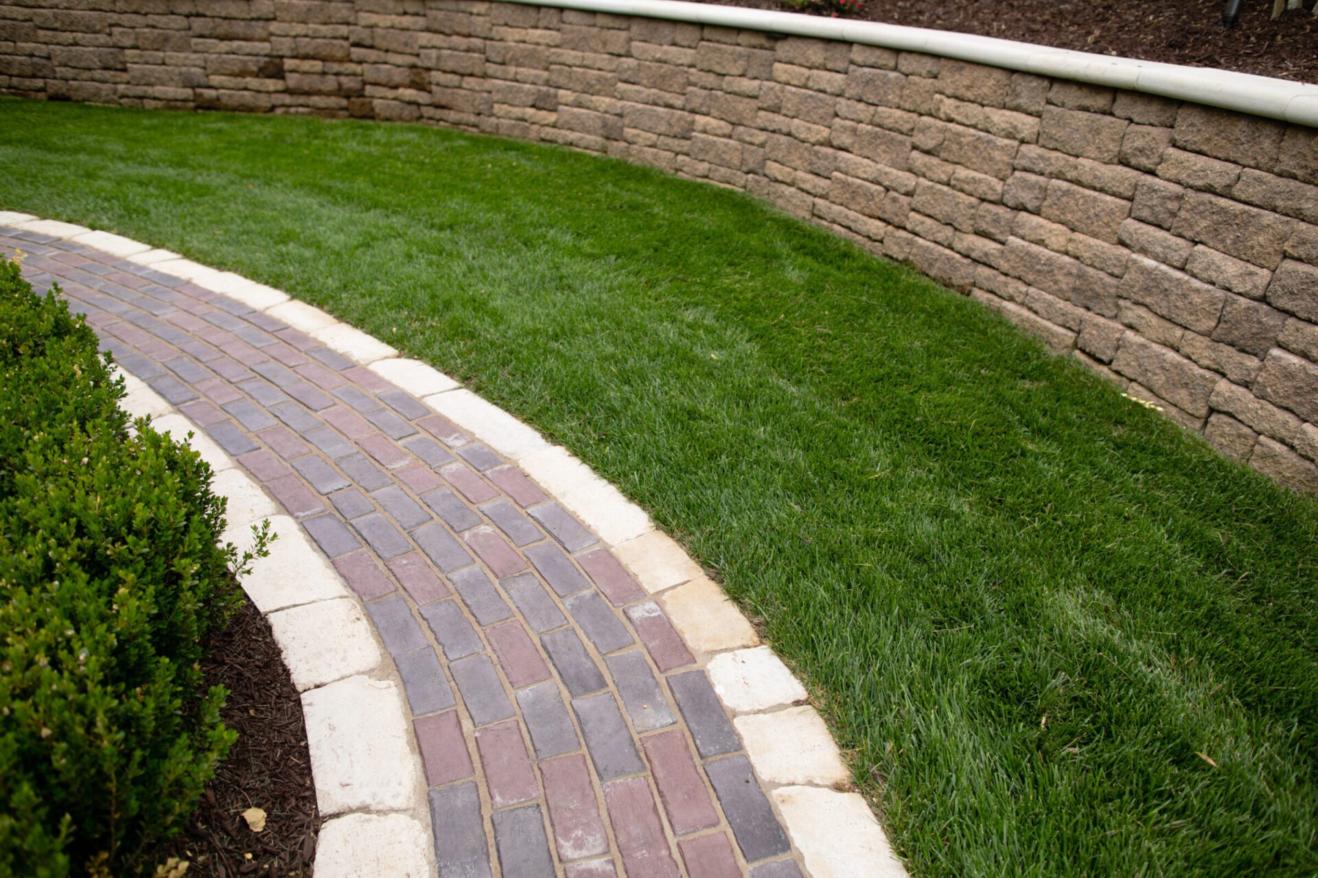 Curved brick path bordered by lush greenery and a stone retaining wall, creating a neat and tidy landscaped garden area.