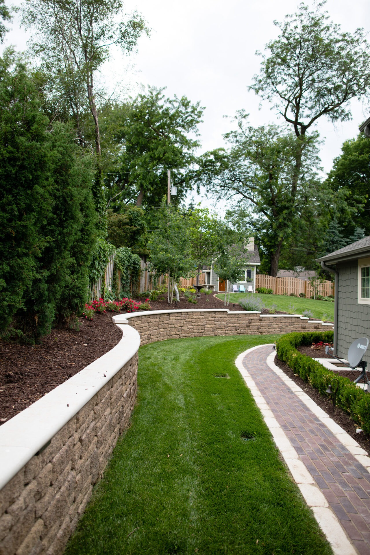 Curved brick walkway and stone retaining wall in landscaped backyard with green grass, trees, and shrubs. Residential home in background.