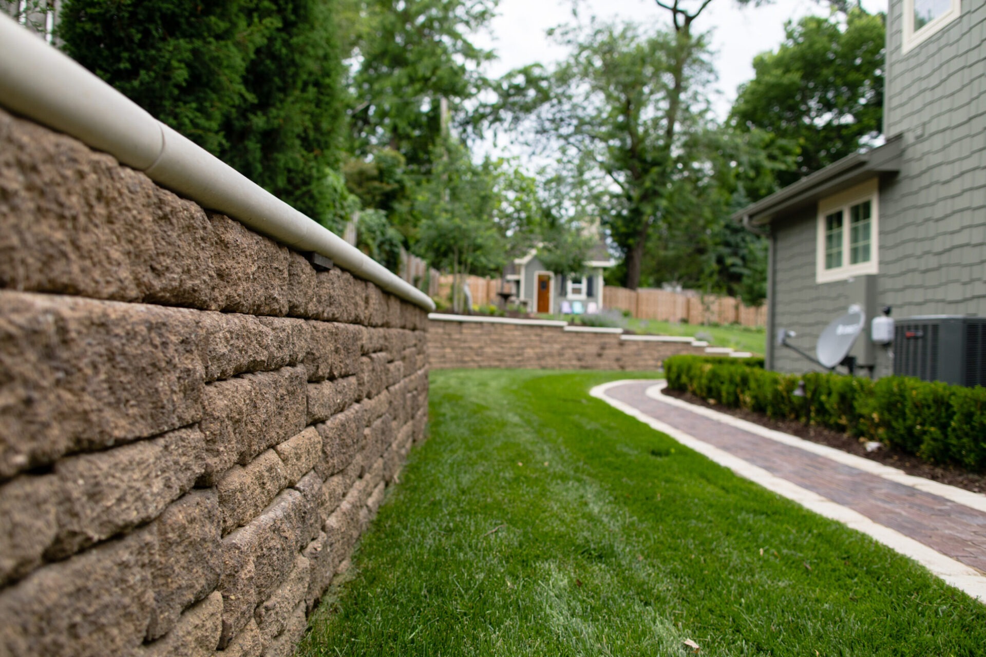 A well-maintained backyard features a stone retaining wall, lush grass, a curved brick path, and a small building in the background.