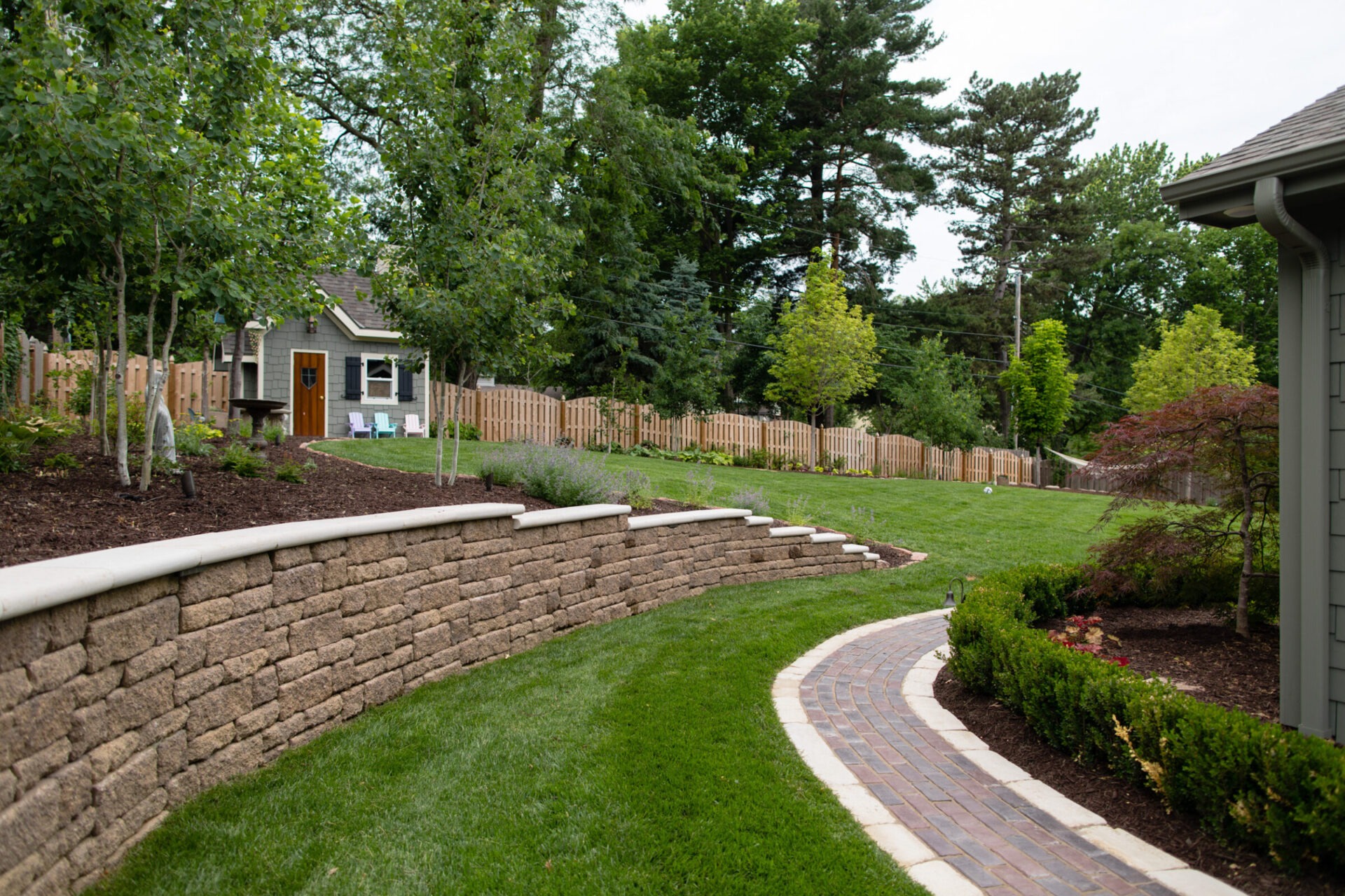 A landscaped backyard features a winding brick path, small shed, manicured lawn, wooden fence, and lush greenery under a clear, bright sky.