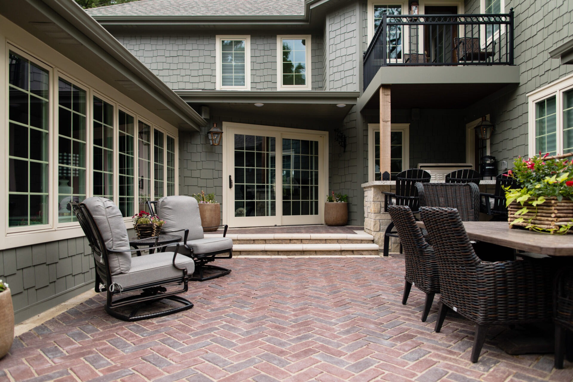 Cozy patio with wicker furniture, plants, and sliding glass doors. Grey shingle house exterior, brick flooring, and a small upper balcony visible.