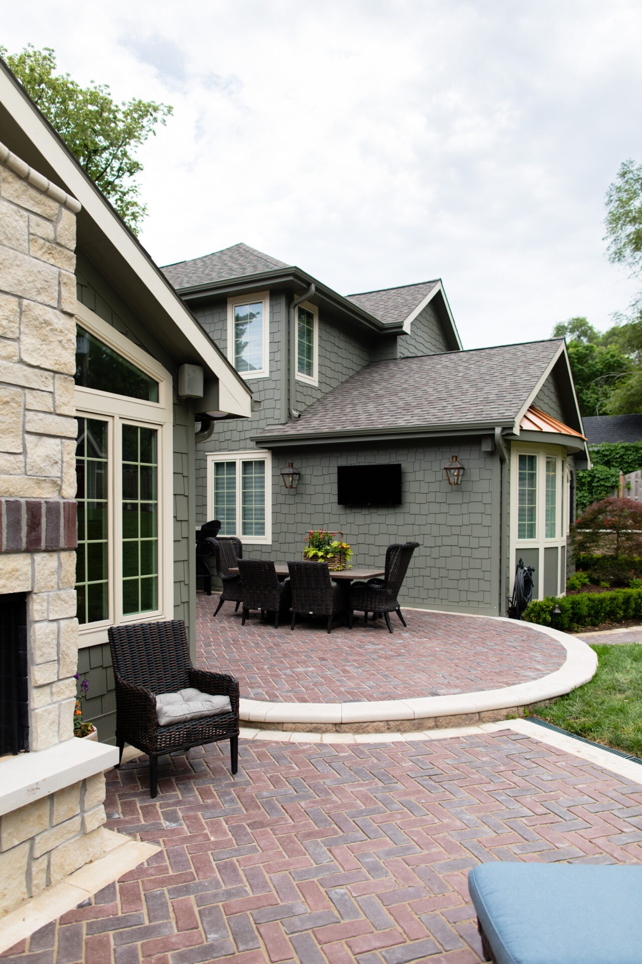 A cozy brick patio with outdoor furniture, a wall-mounted TV, and a person near a grill at a modern house.