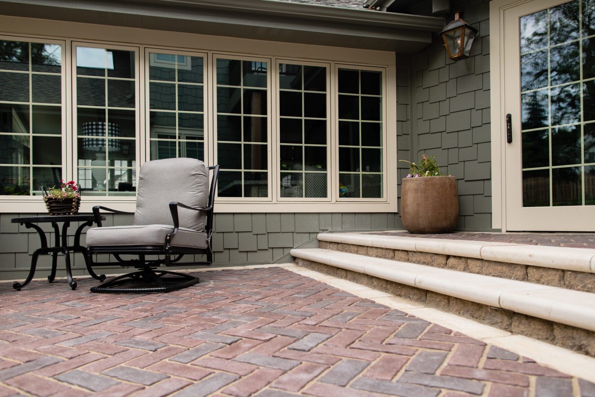 Cozy patio facade with cushioned chair, small table, and potted plant on a brick walkway, leading to windows and door entryway.