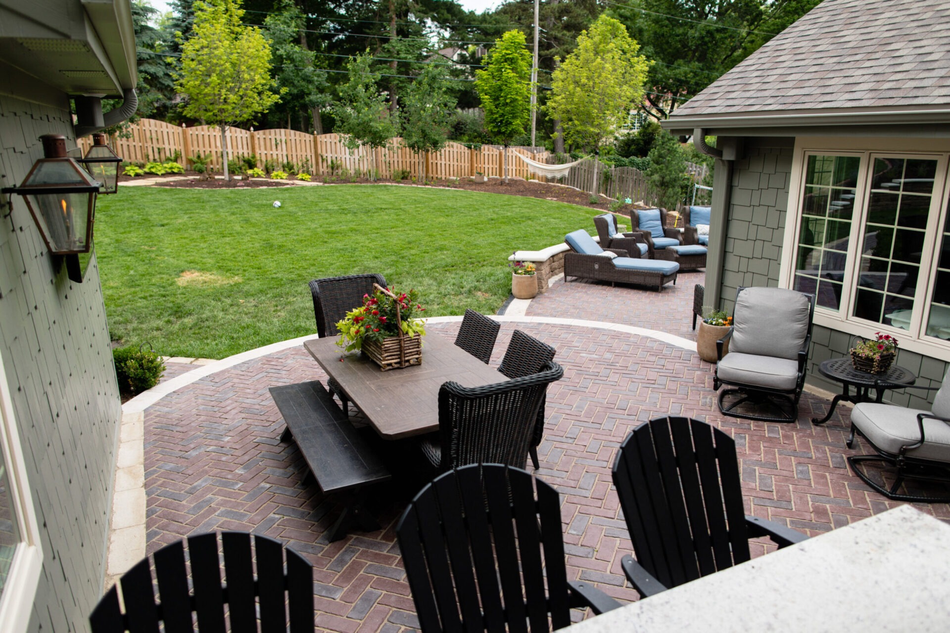 A cozy backyard with a brick patio area, featuring outdoor seating, a dining table, and a green lawn surrounded by a wooden fence.