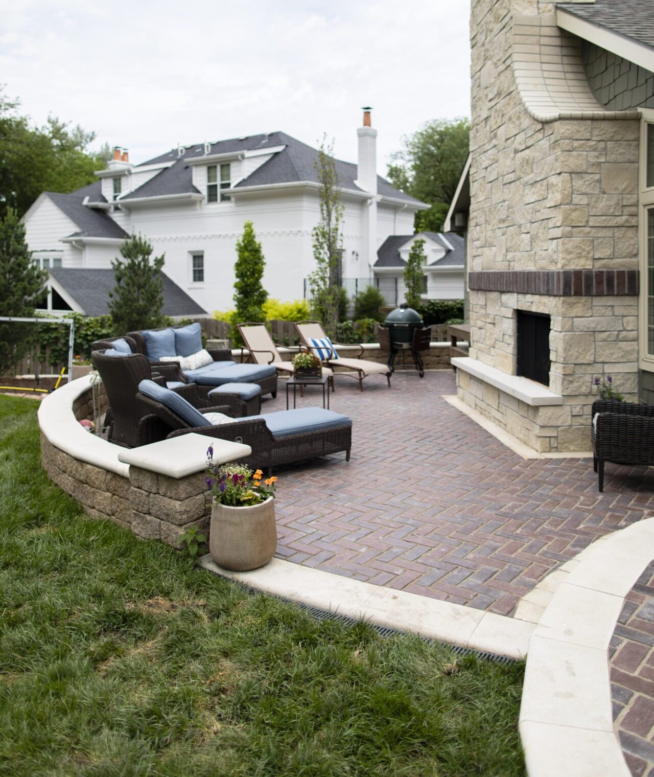 A spacious patio showing off one of our clients' outdoor kitches which features elegant outdoor seating, stone fireplace, and potted plants, with a large, stylish house in the background amidst greenery.