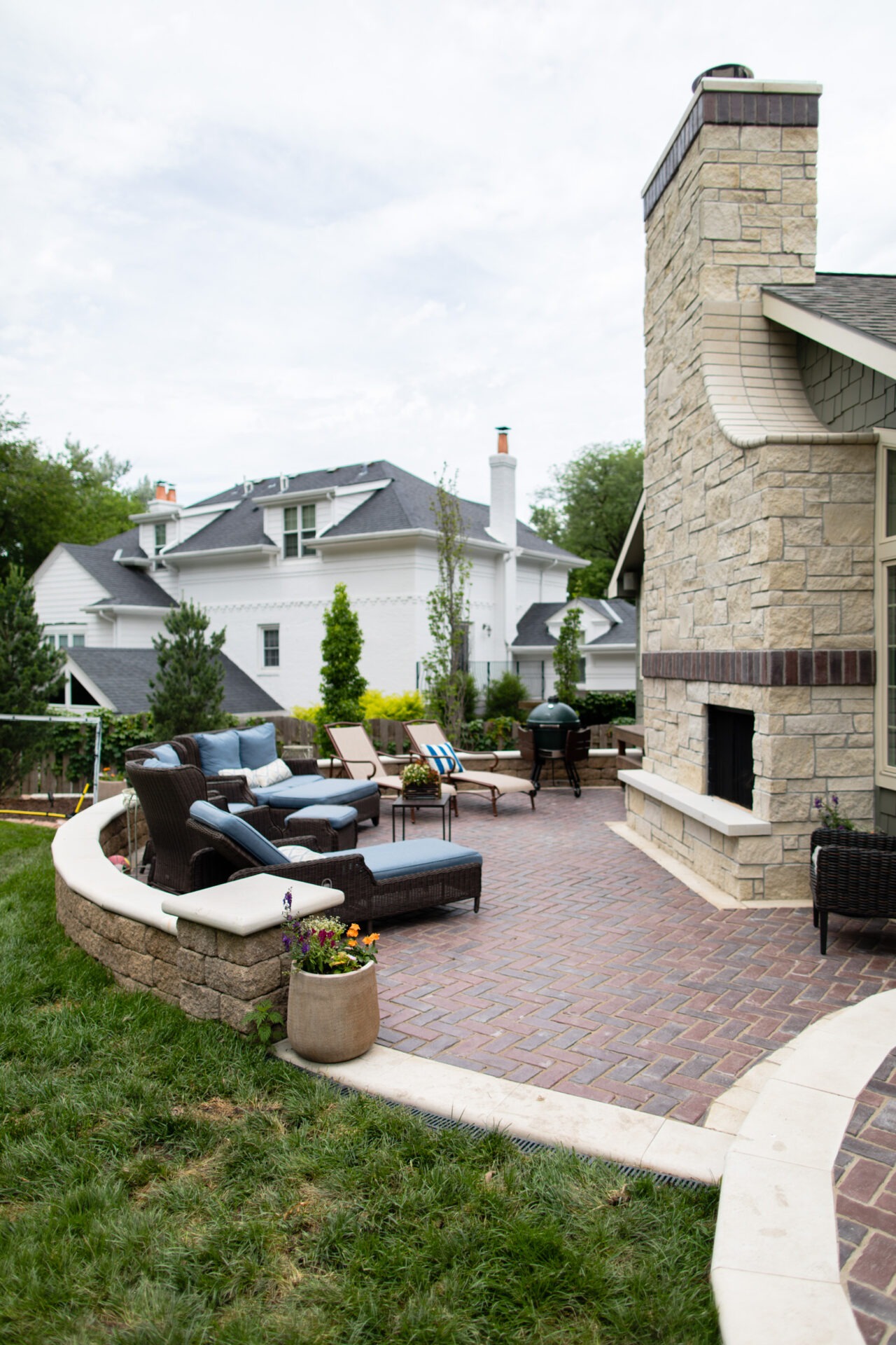 Spacious patio with wicker seating, stone fireplace, and landscaped garden, adjoining a large white house. Relaxing outdoor setting in a residential area.