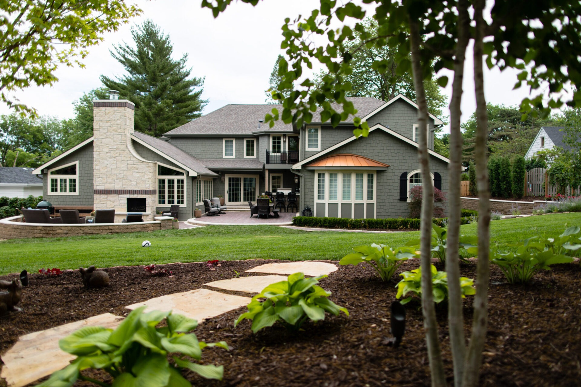 A large, modern house with a landscaped garden, patio area, and outdoor fireplace, surrounded by trees and well-maintained lawn.