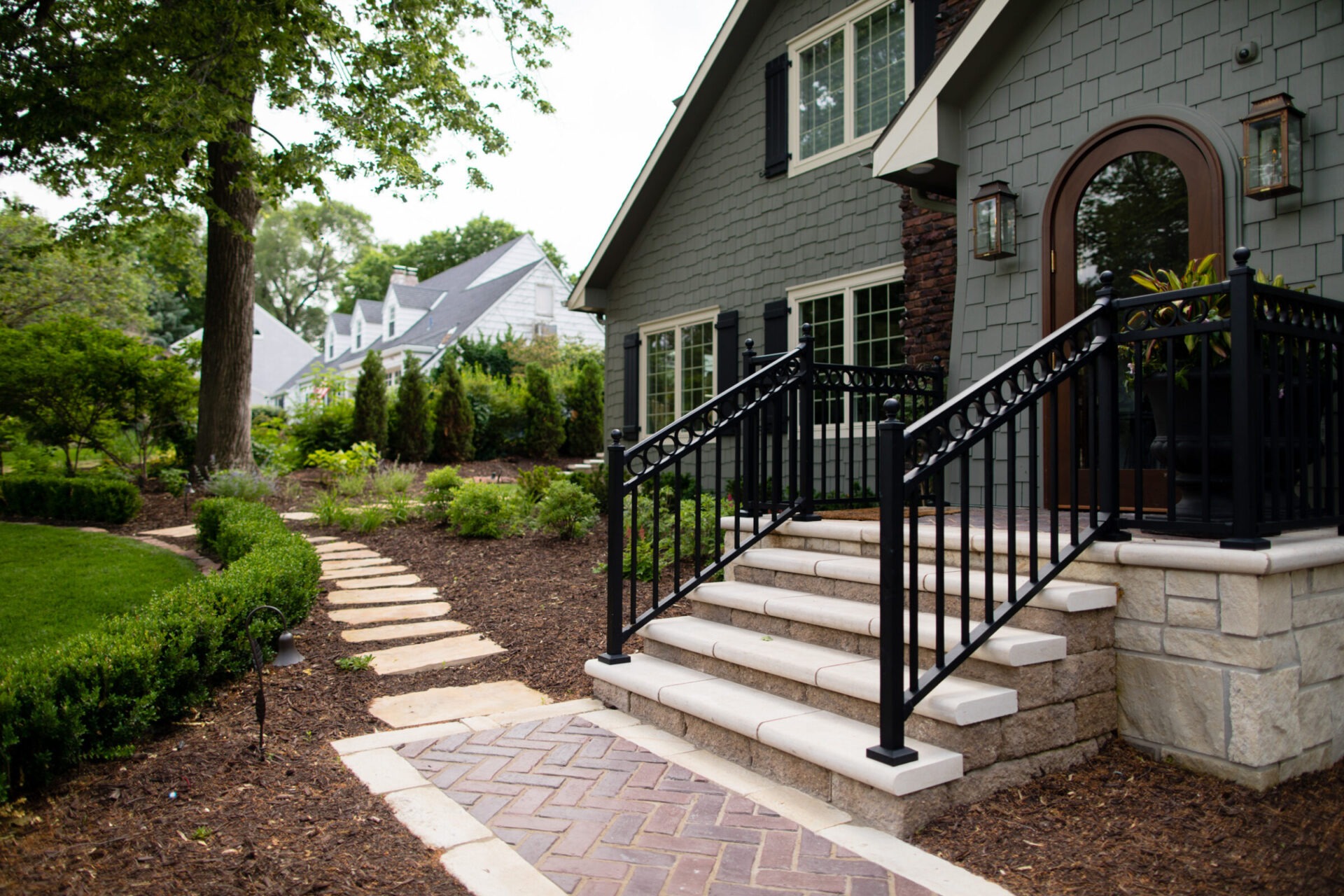 A charming house with a stone path, surrounded by lush greenery and landscaped gardens, featuring a black-railed staircase leading to the entrance.