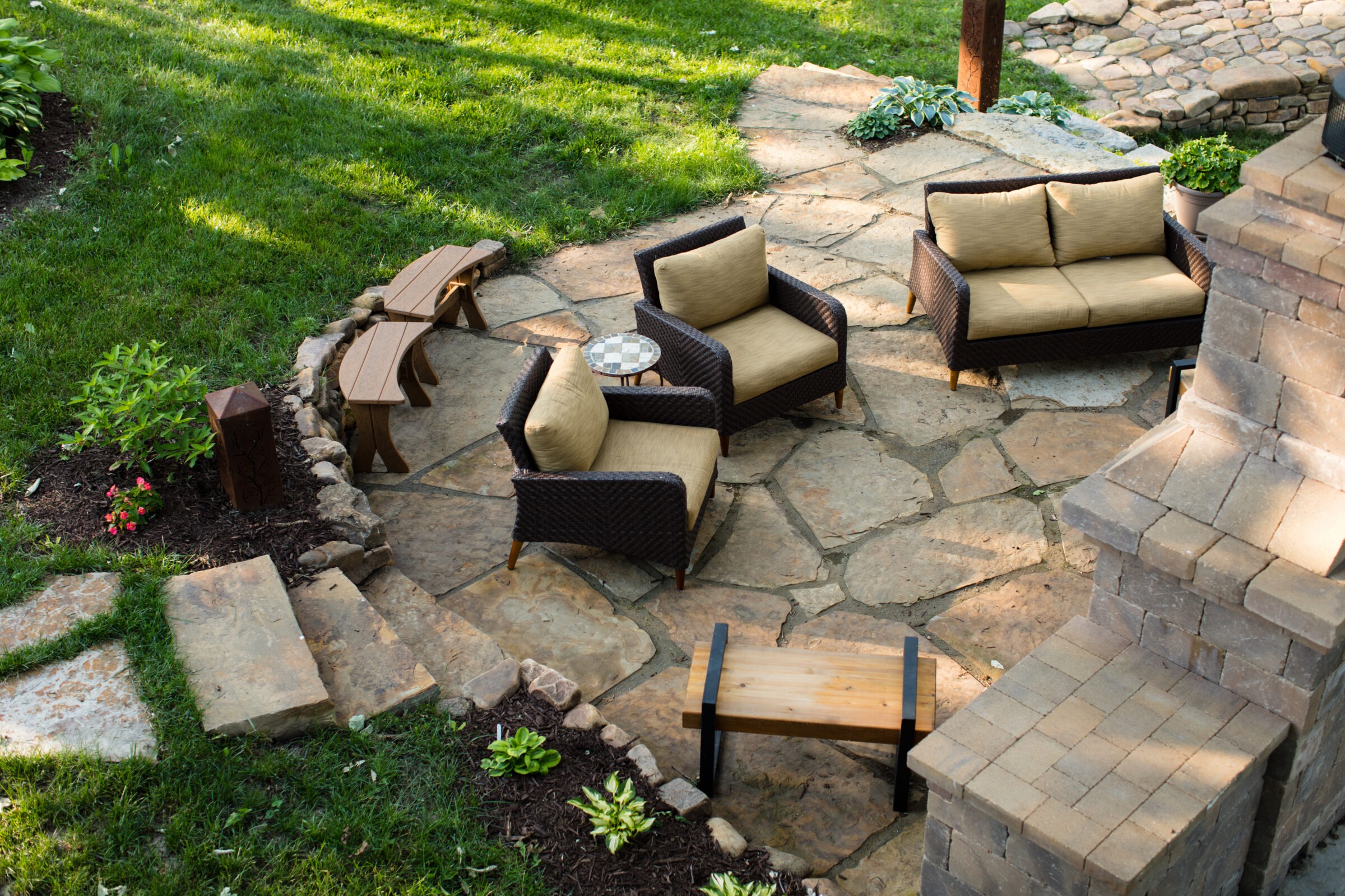 Stone patio with wicker furniture, surrounded by green grass and plants. Includes a brick structure, wooden benches, creating a cozy outdoor space.