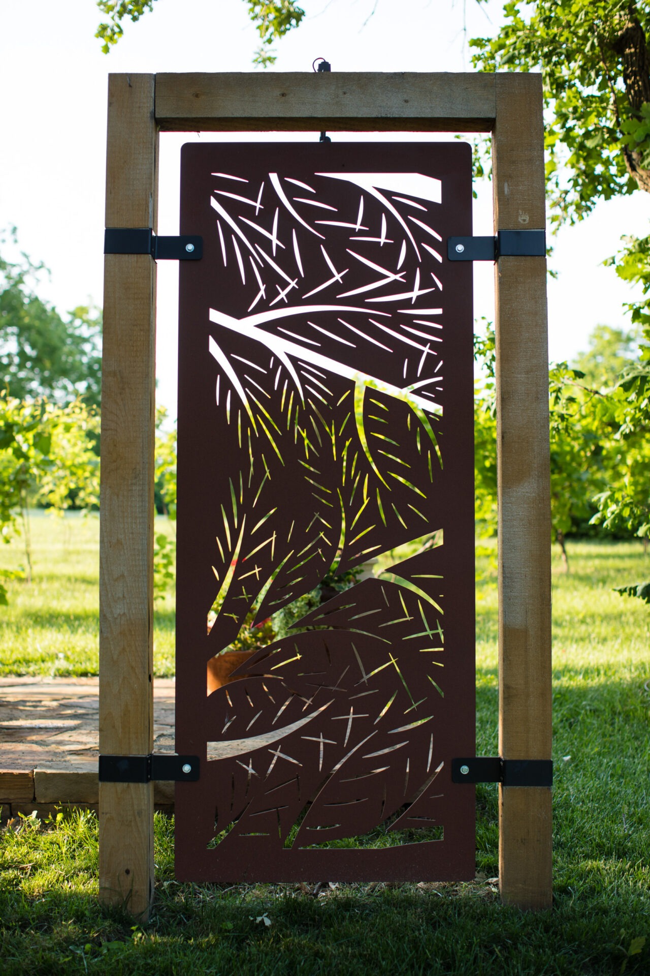 Decorative metal gate with leaf pattern in wooden frame, set in a lush green garden, surrounded by trees and sunshine filtering through.