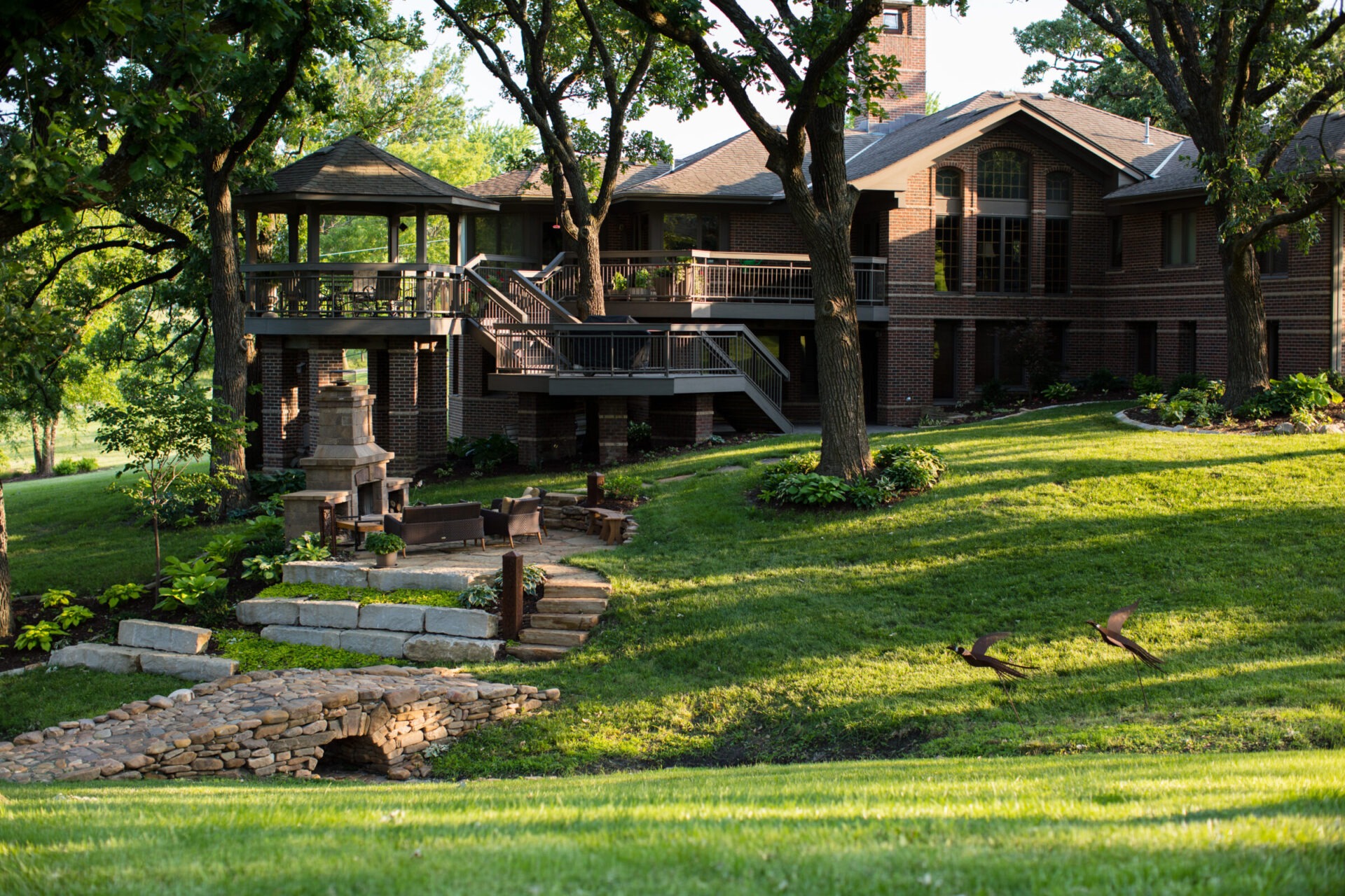 A brick house with two stories features a spacious patio, outdoor fireplace, lush lawn, trees, and a small stone footbridge.