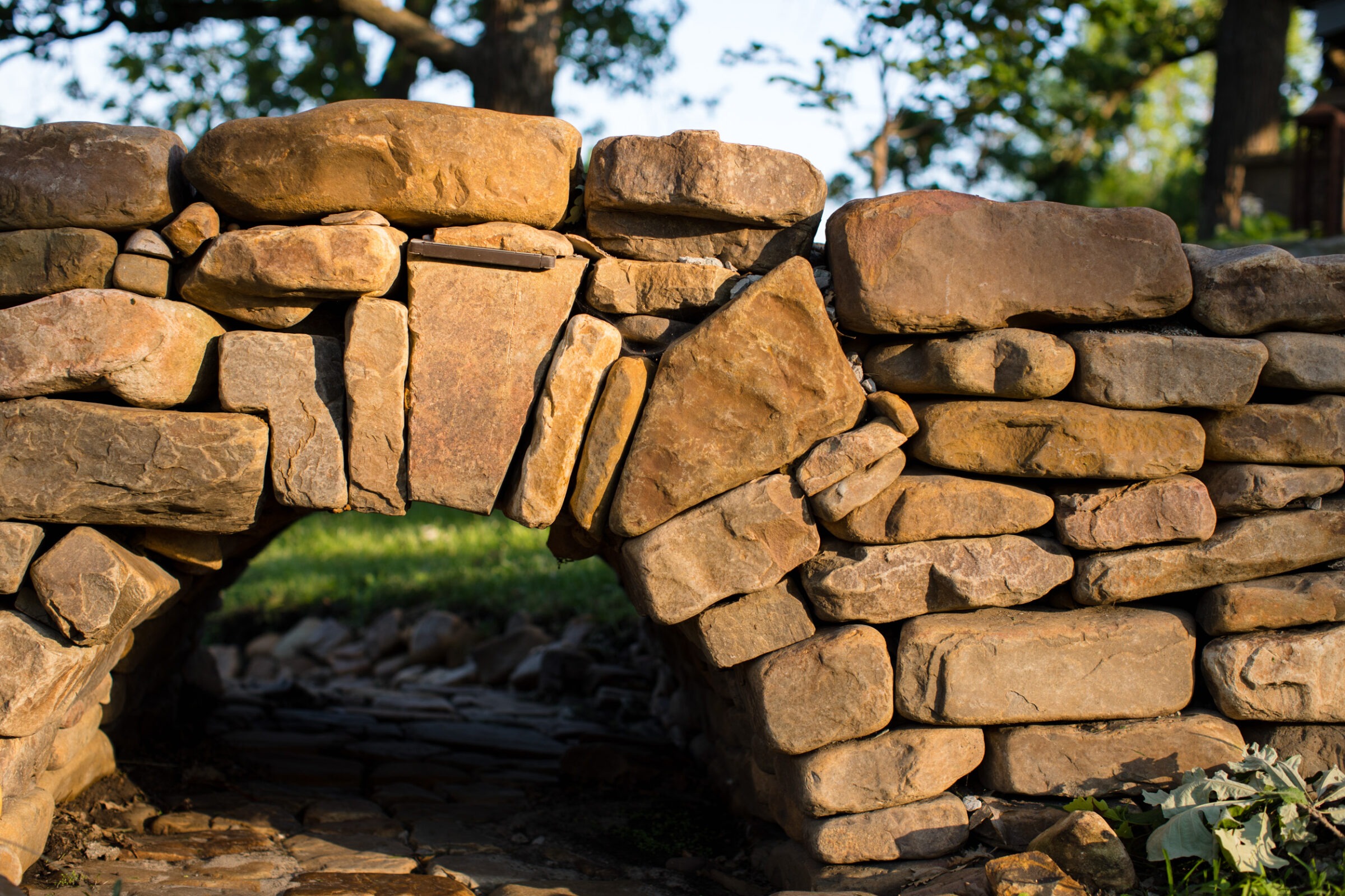 Sunlit stone archway constructed from rough stones, with trees and grass visible in the background, creating a rustic, natural atmosphere.