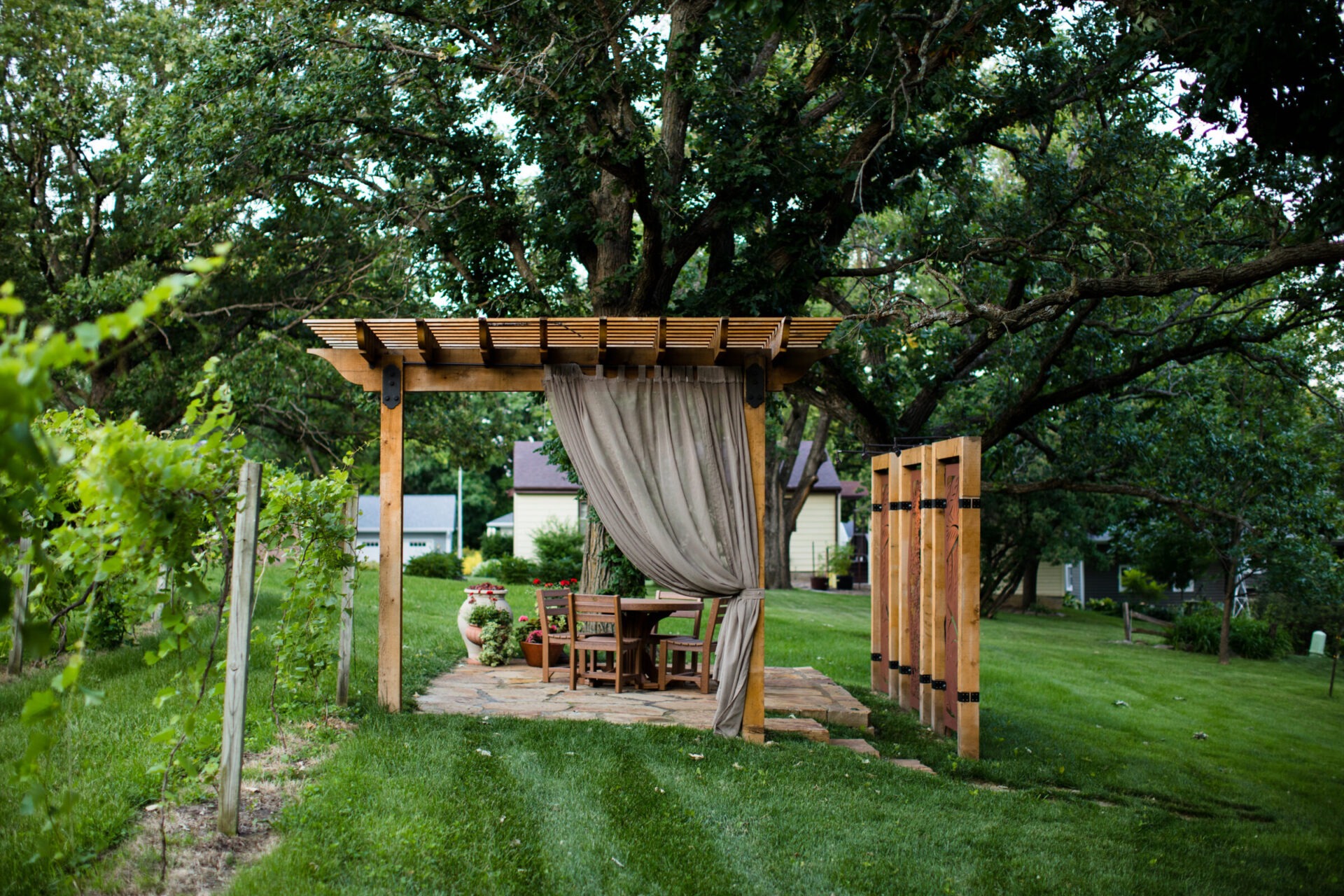 A wooden pergola with curtains and a table set in a lush garden surrounded by vineyards and trees, creating a serene atmosphere.