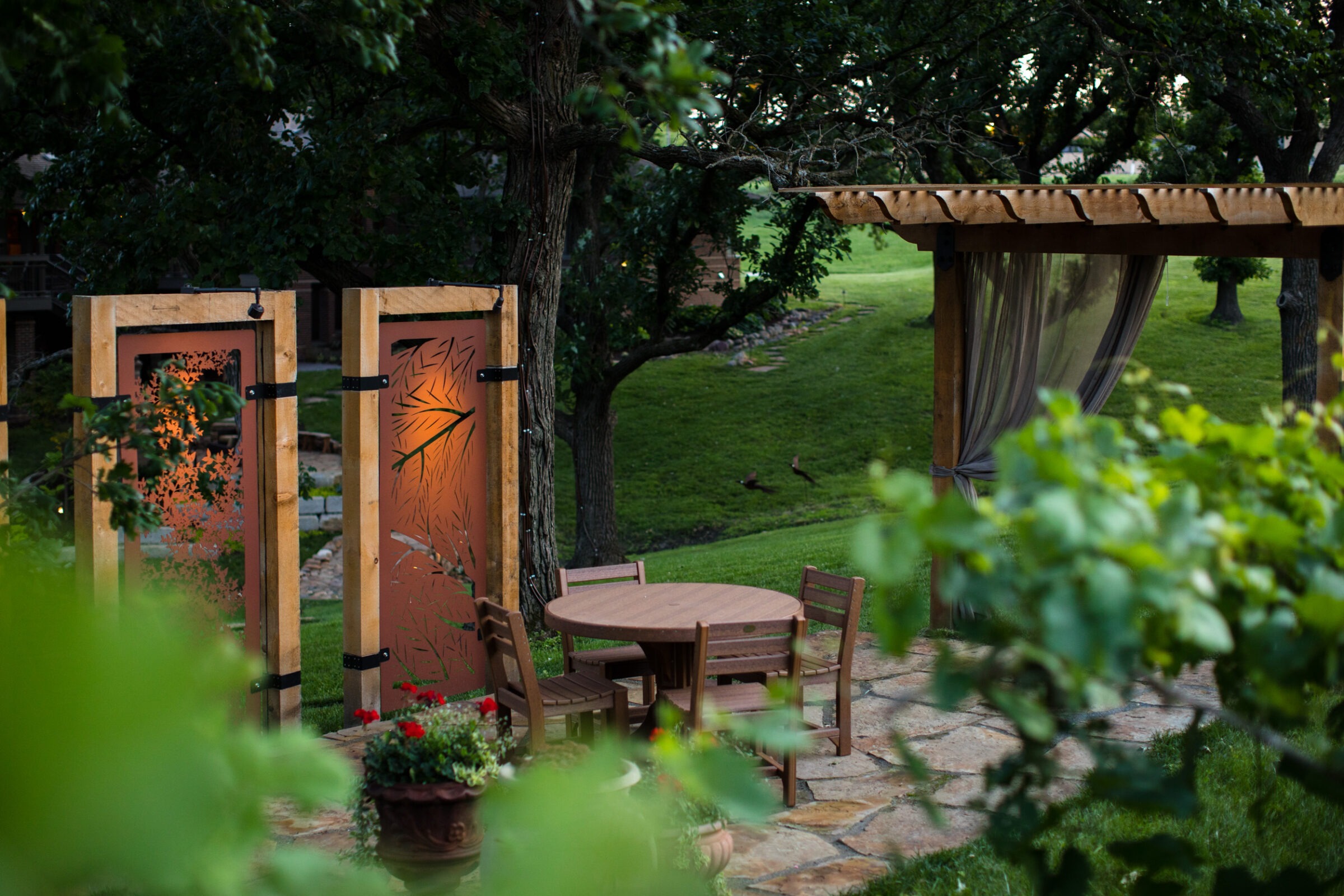 A serene garden features a wooden pergola, a round table with chairs, decorative screens, lush greenery, and trees enhancing the tranquil atmosphere.