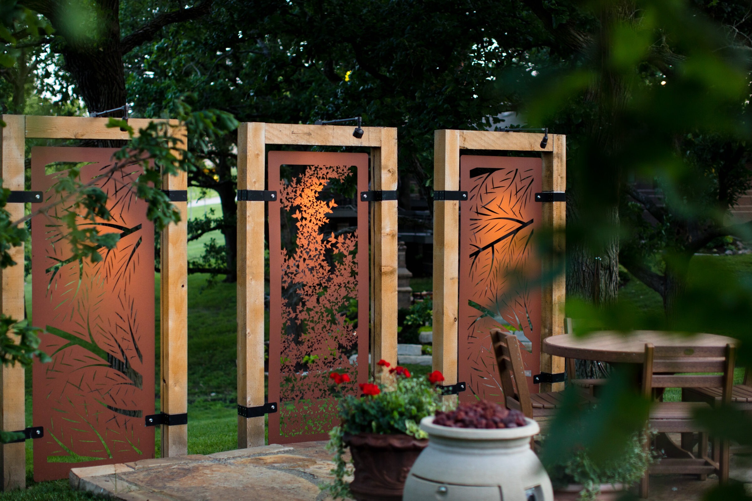 Decorative metal screens with leaf patterns in a garden setting. Wooden round table, chairs, and potted plants complete the peaceful outdoor space.