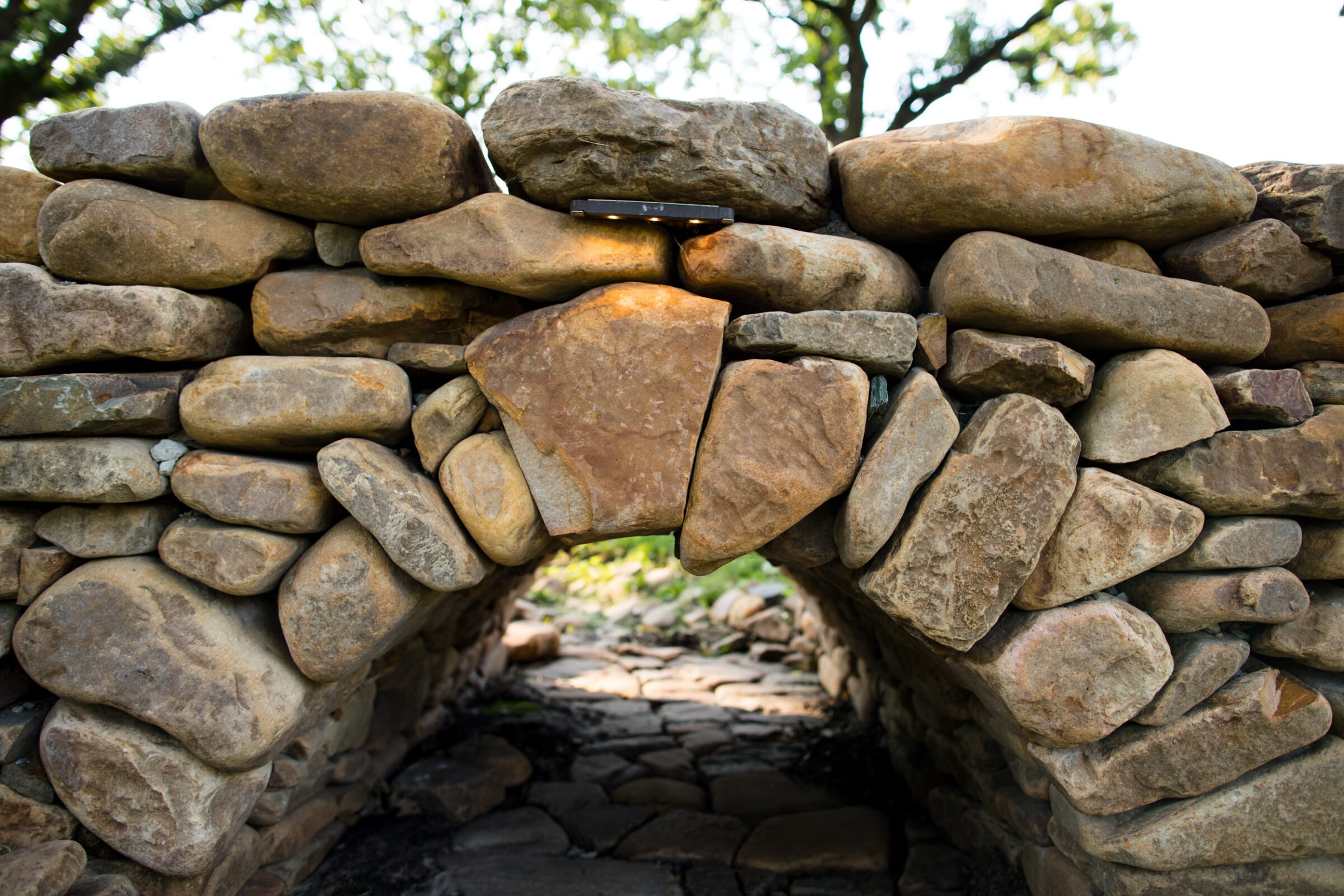 Stone archway built with large rocks, showing a natural landscape background with trees. Simple, rustic design with natural materials.