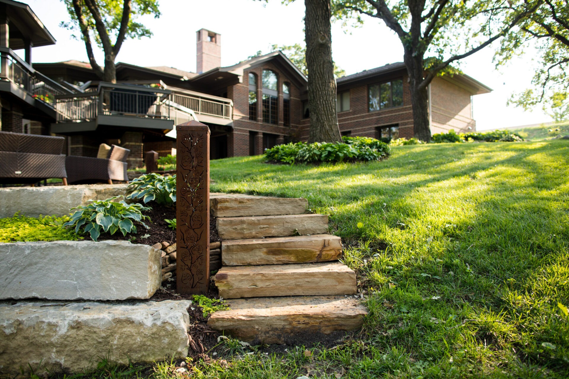 Stone steps lead to a landscaped yard with shrubs and trees beside a large, modern brick house. Cozy outdoor furniture is visible.